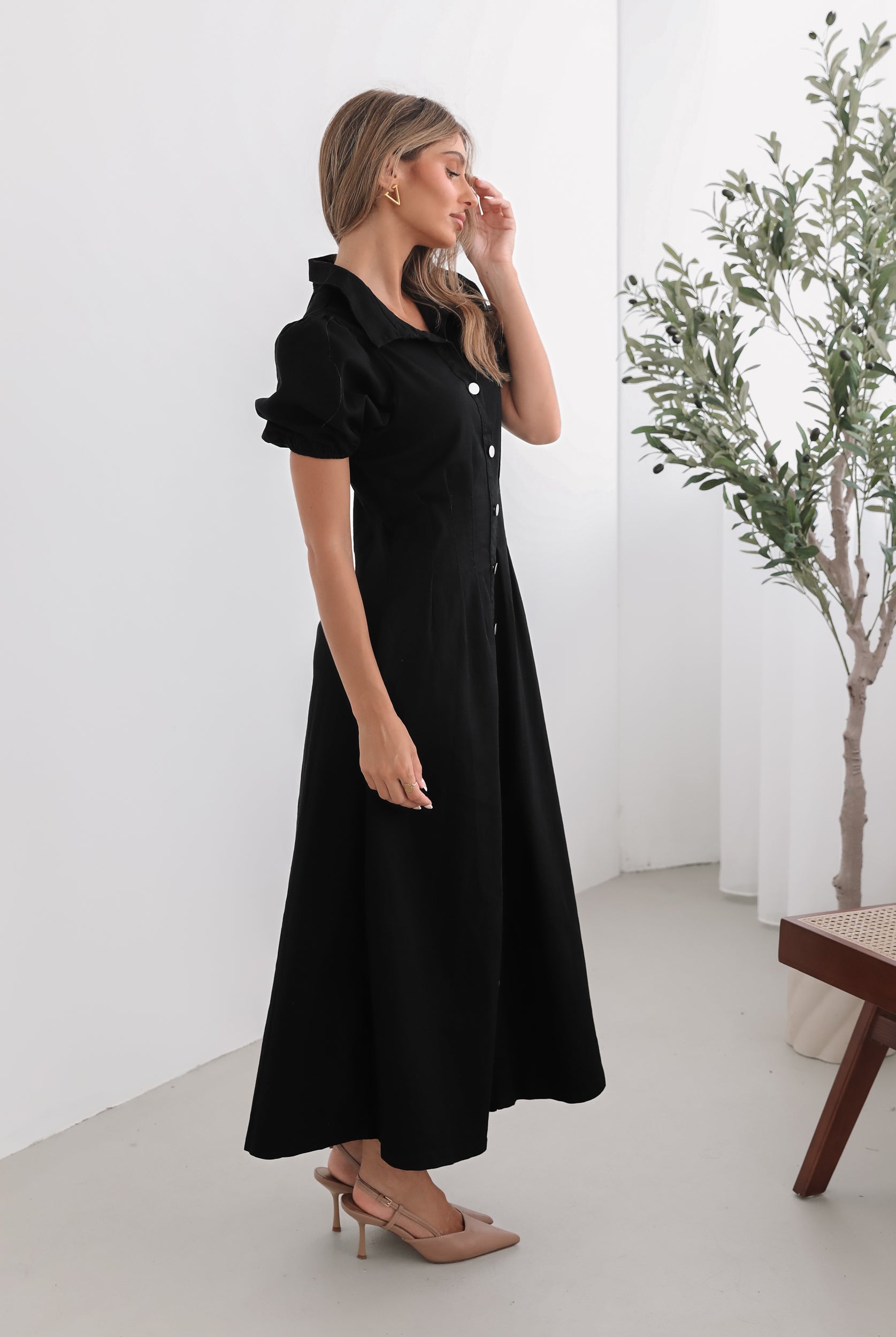 A woman standing in a room with a white background wearing a black knee-length dress with a shirt collar, short puff sleeves, and center-front buttons.