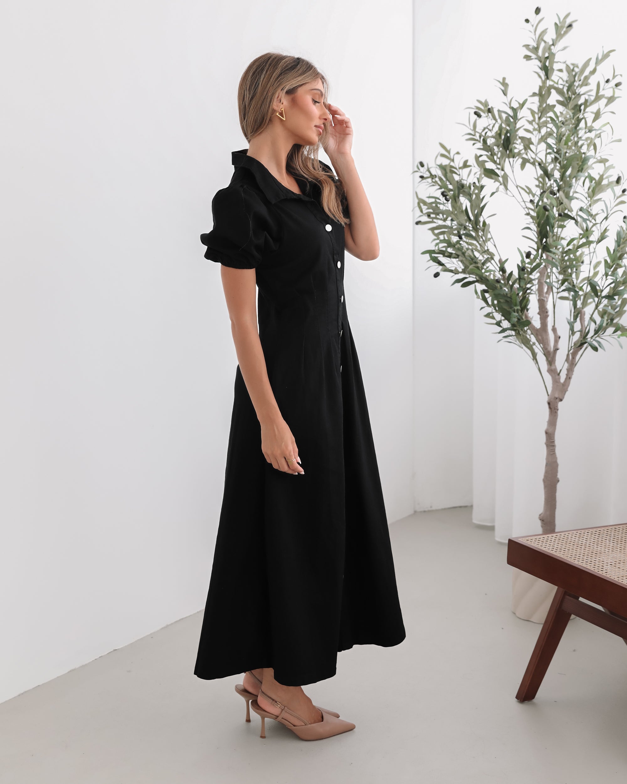 A woman standing in a room with a white background wearing a black knee-length dress with a shirt collar, short puff sleeves, and center-front buttons.