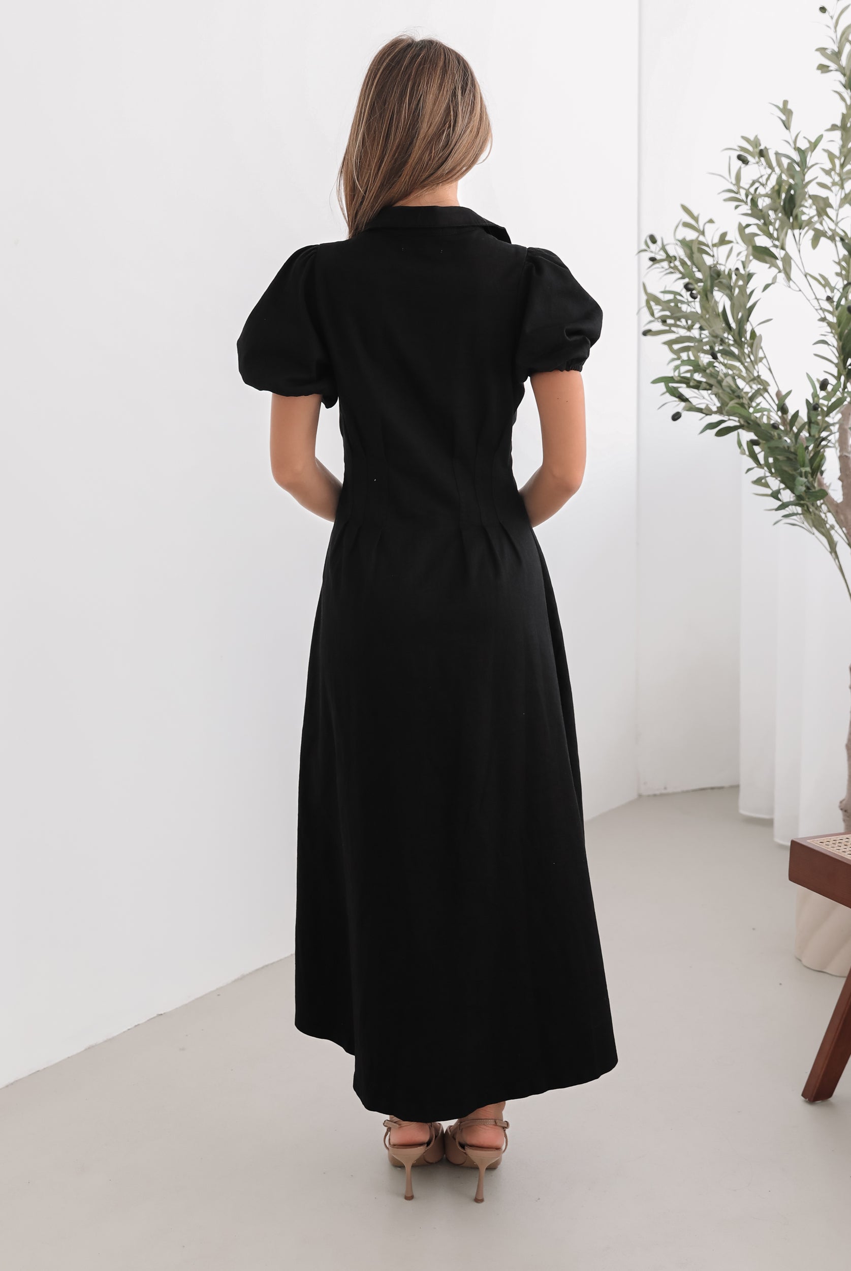 A woman standing in a room with a white background wearing a black knee-length dress with a shirt collar, short puff sleeves, and center-front buttons.