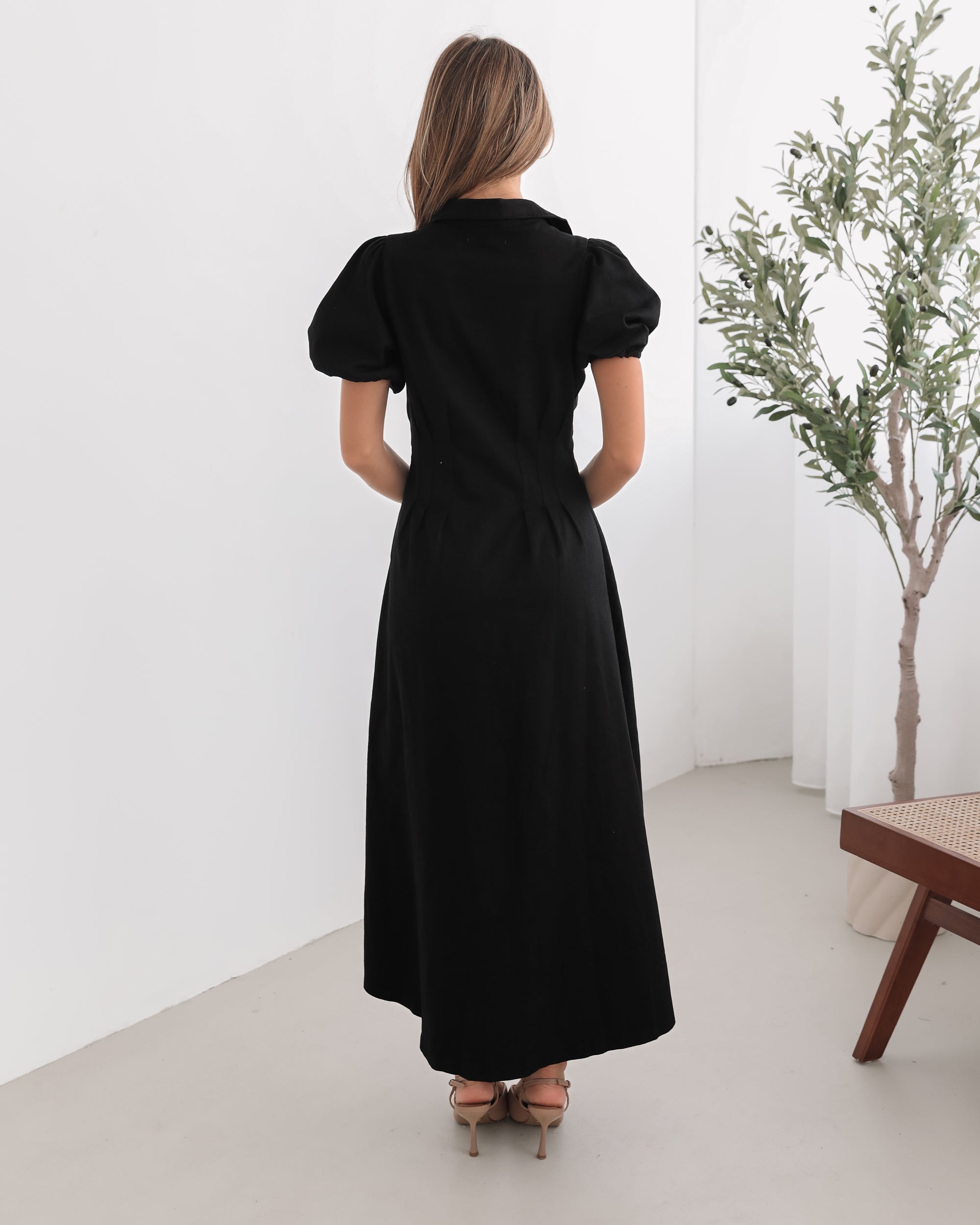 A woman standing in a room with a white background wearing a black knee-length dress with a shirt collar, short puff sleeves, and center-front buttons.