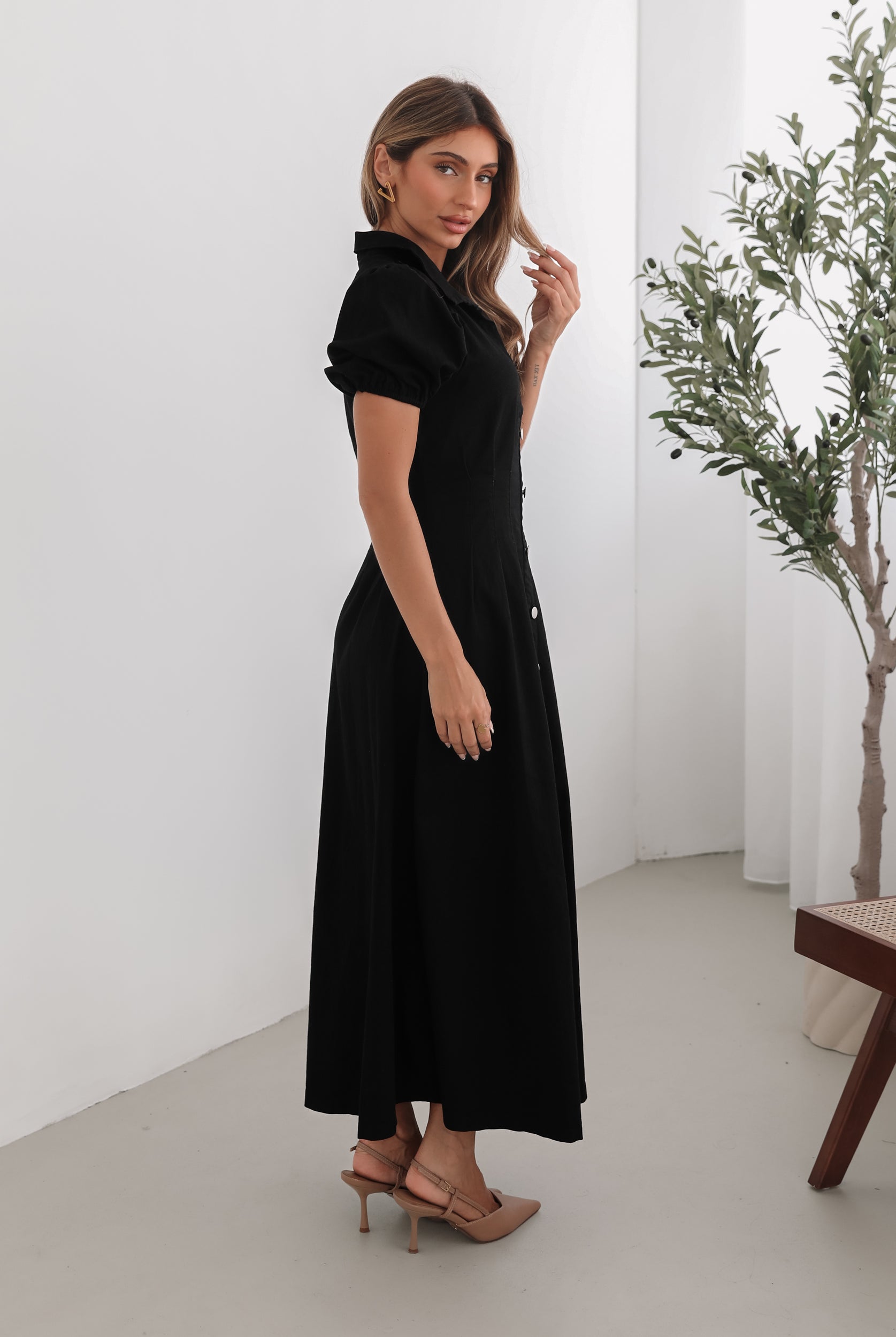 A woman standing in a room with a white background wearing a black knee-length dress with a shirt collar, short puff sleeves, and center-front buttons.