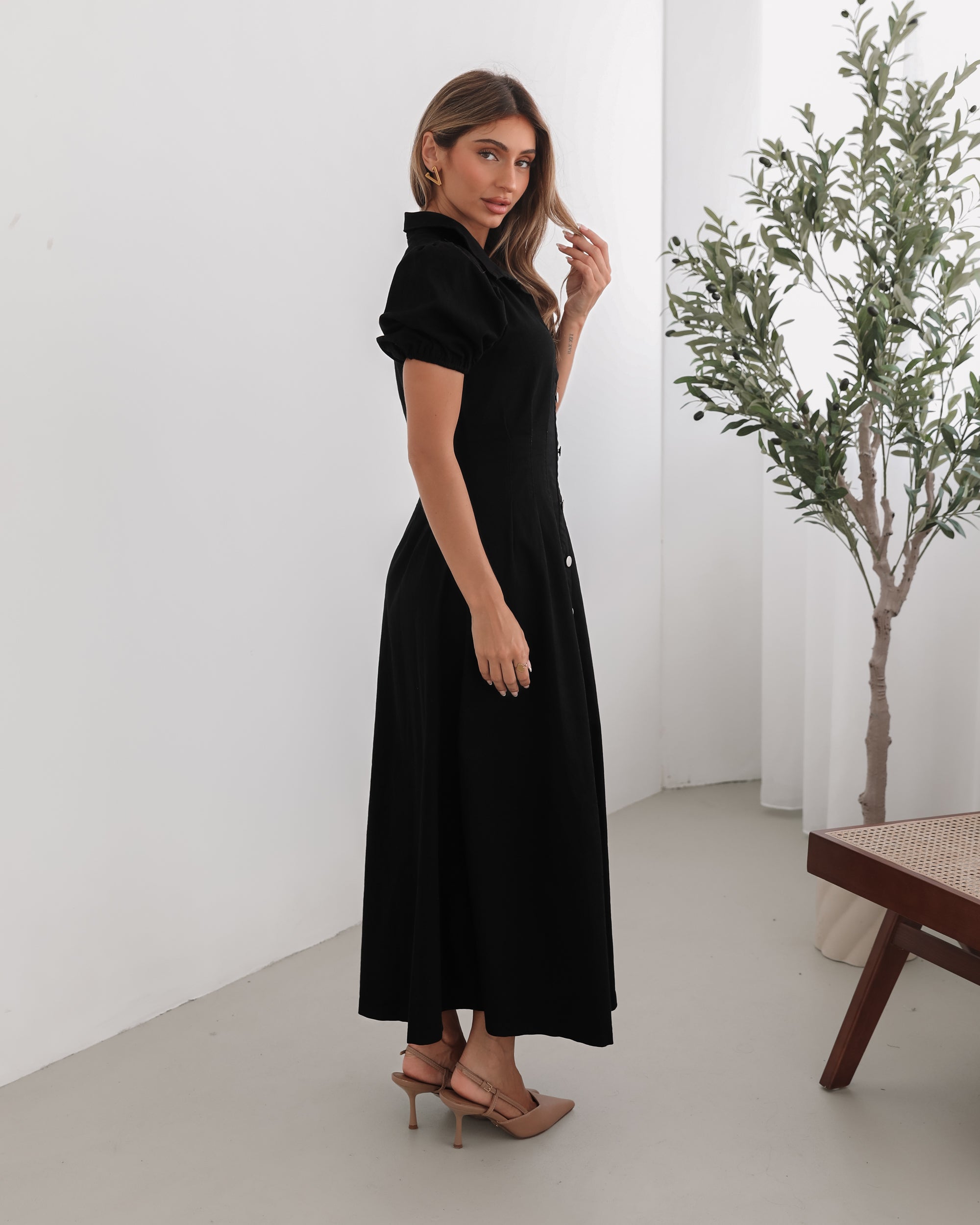 A woman standing in a room with a white background wearing a black knee-length dress with a shirt collar, short puff sleeves, and center-front buttons.