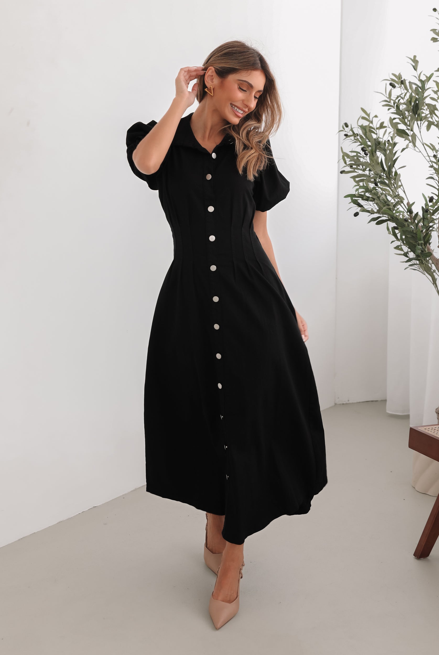 A woman standing in a room with a white background wearing a black knee-length dress with a shirt collar, short puff sleeves, and center-front buttons.