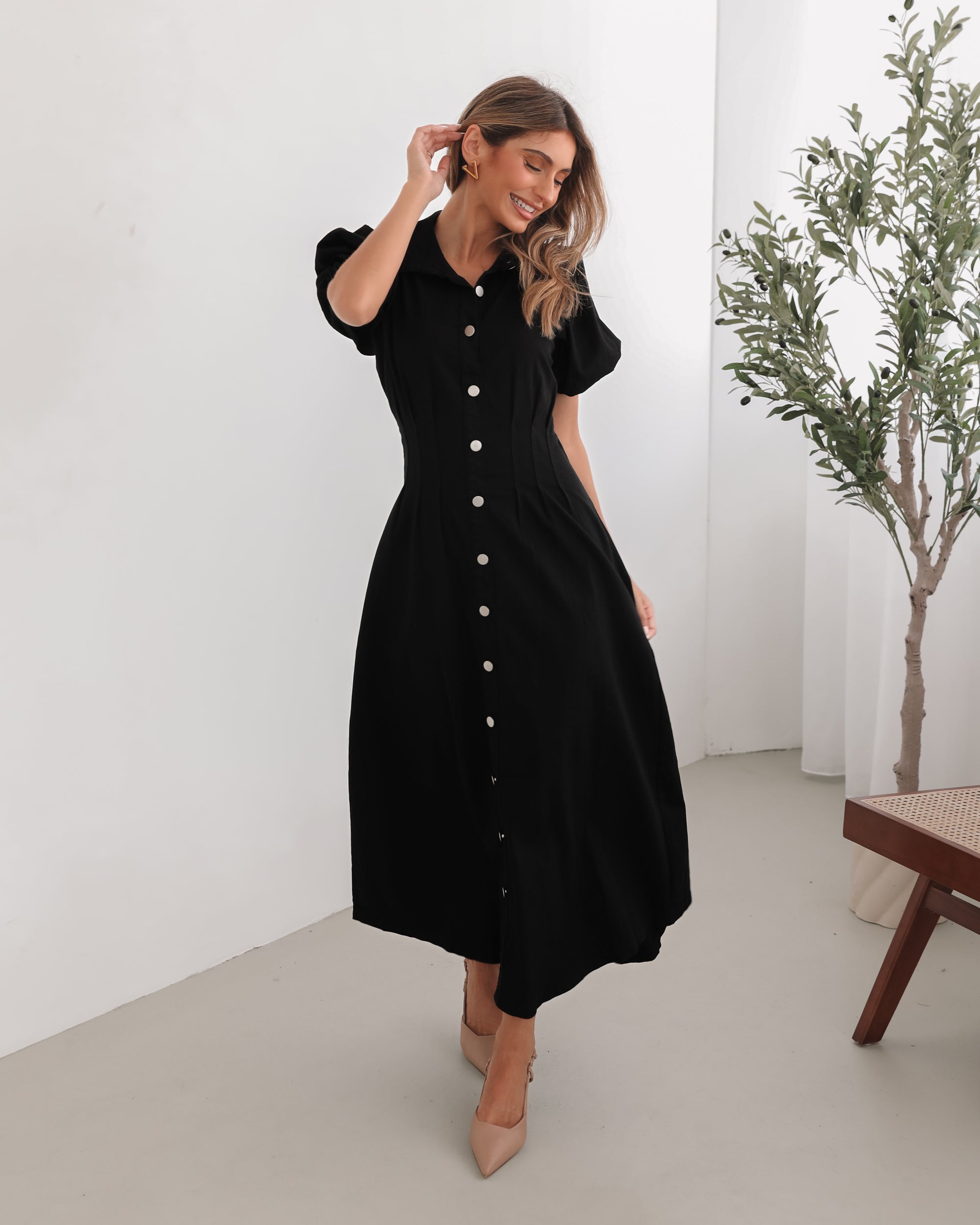 A woman standing in a room with a white background wearing a black knee-length dress with a shirt collar, short puff sleeves, and center-front buttons.