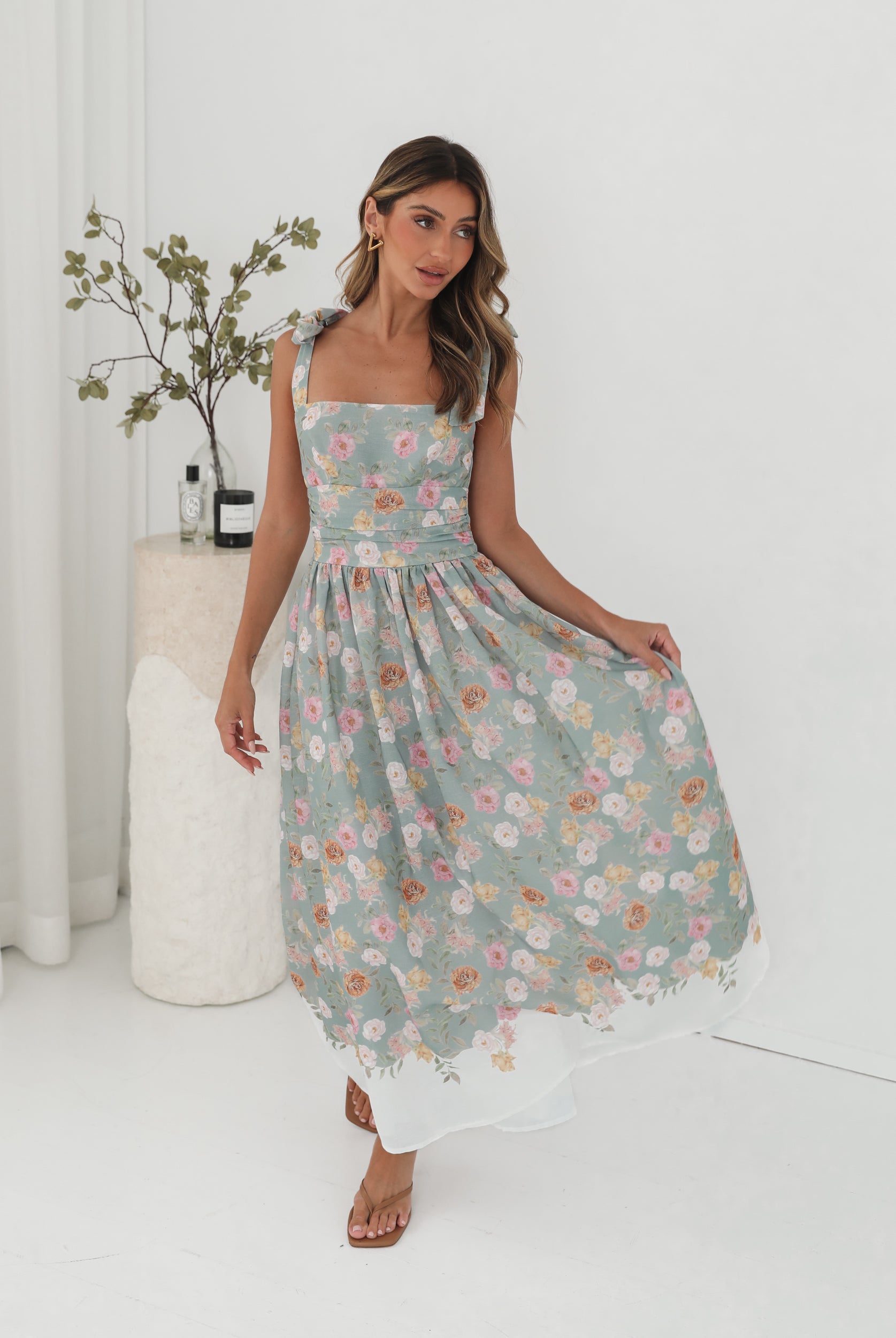 A woman standing in a room with a white background, wearing a mint floral dress with bow straps and a square neckline.