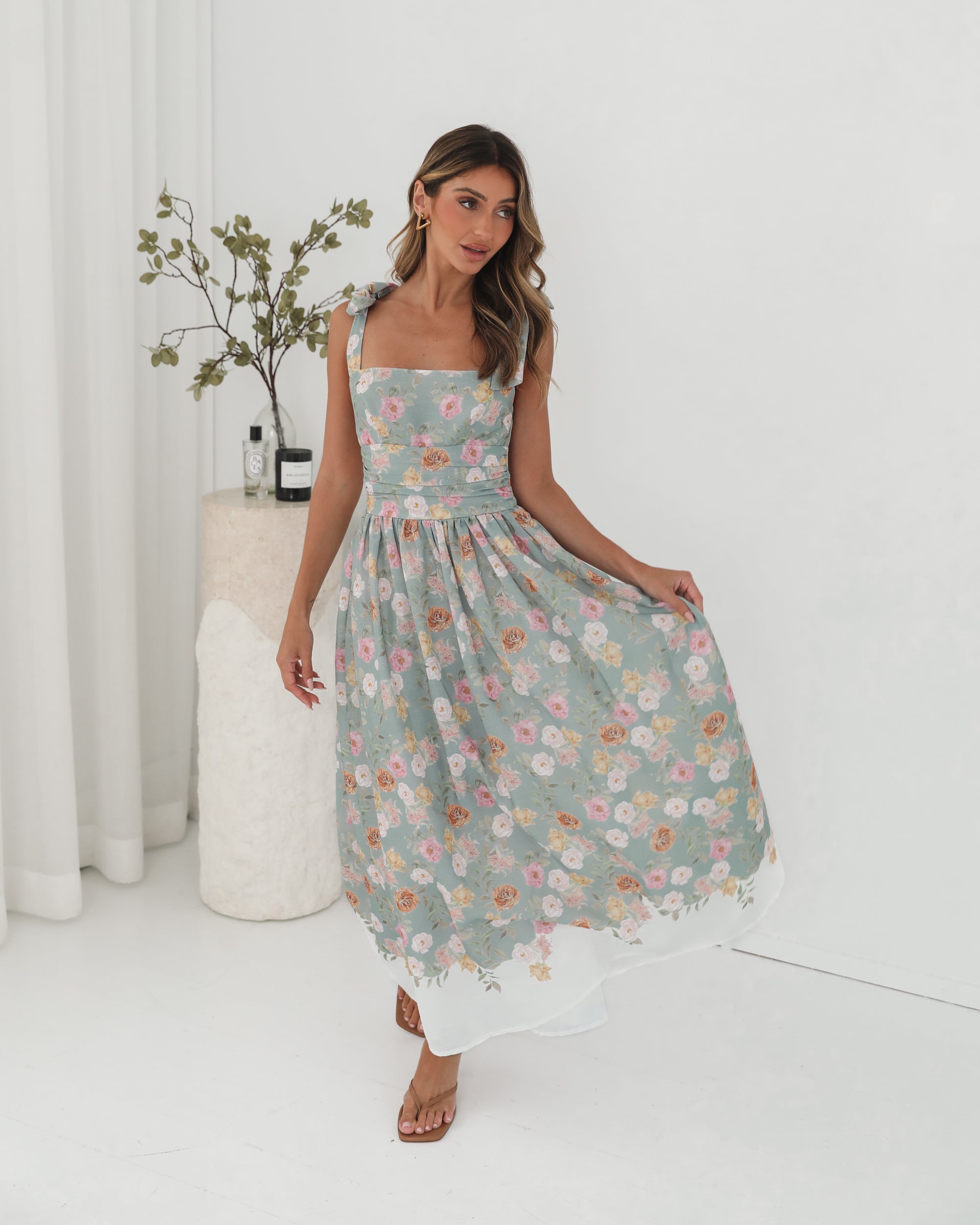 A woman standing in a room with a white background, wearing a mint floral dress with bow straps and a square neckline.