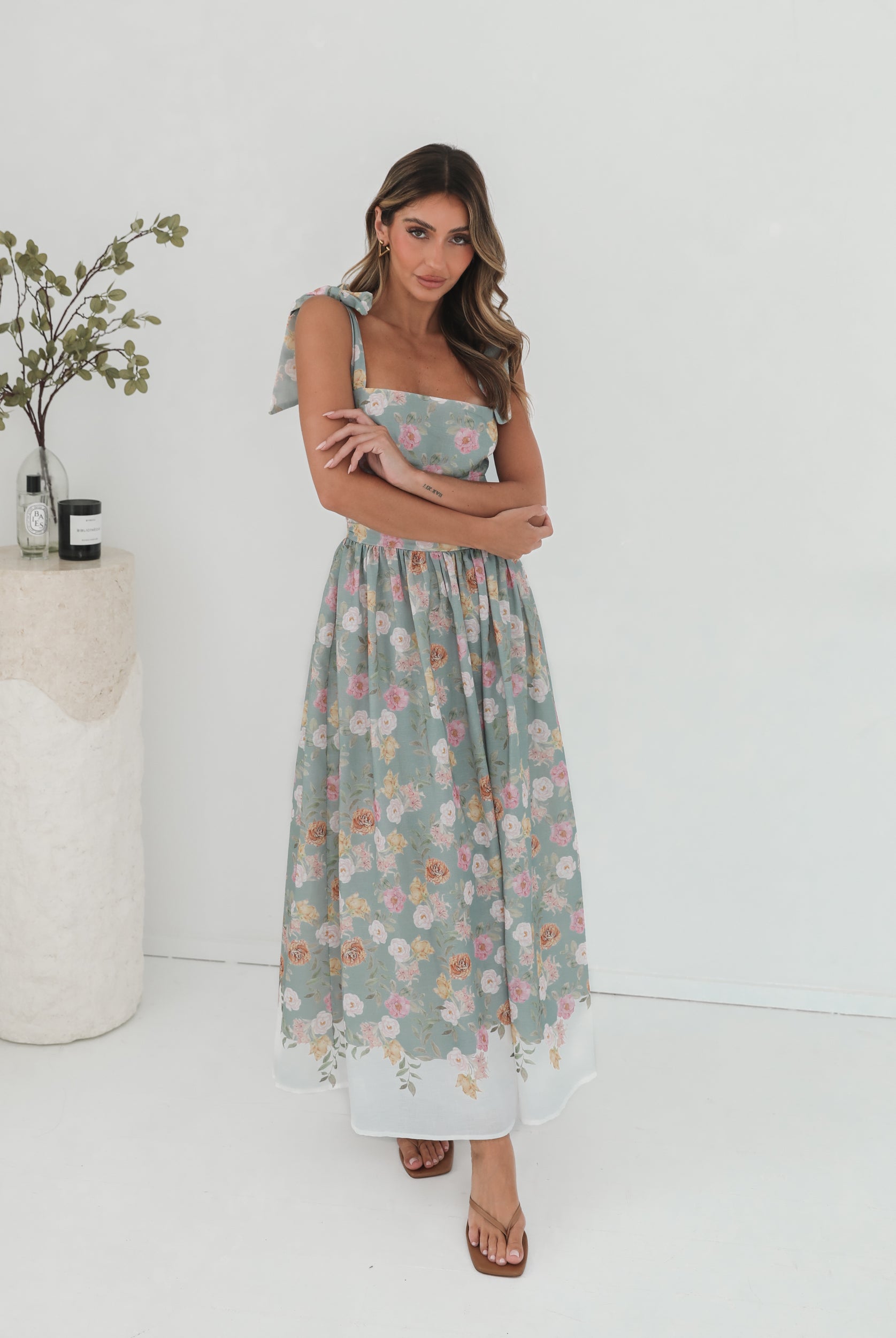 A woman standing in a room with a white background, wearing a mint floral dress with bow straps and a square neckline.