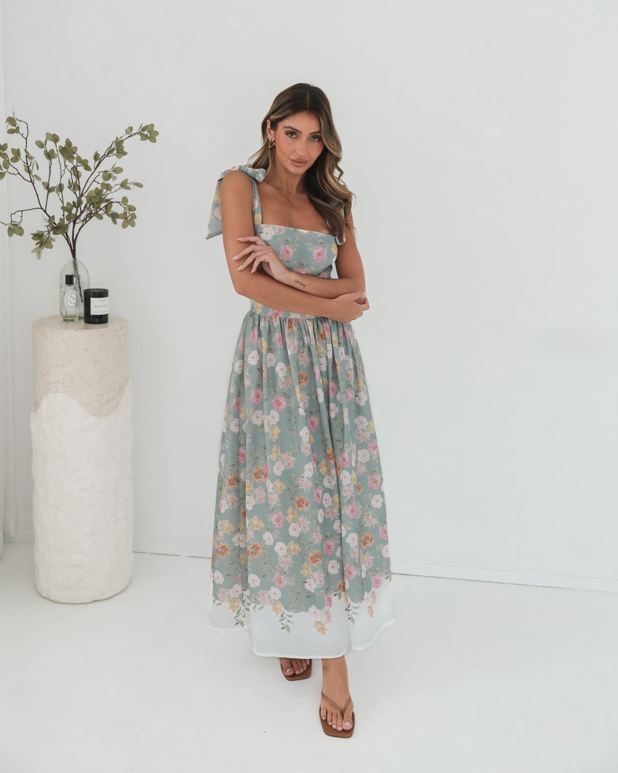A woman standing in a room with a white background, wearing a mint floral dress with bow straps and a square neckline.