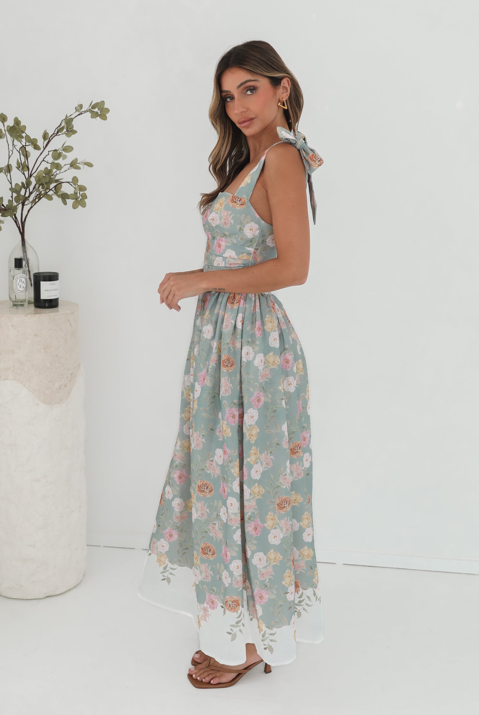 A woman standing in a room with a white background, wearing a mint floral dress with bow straps and a square neckline.