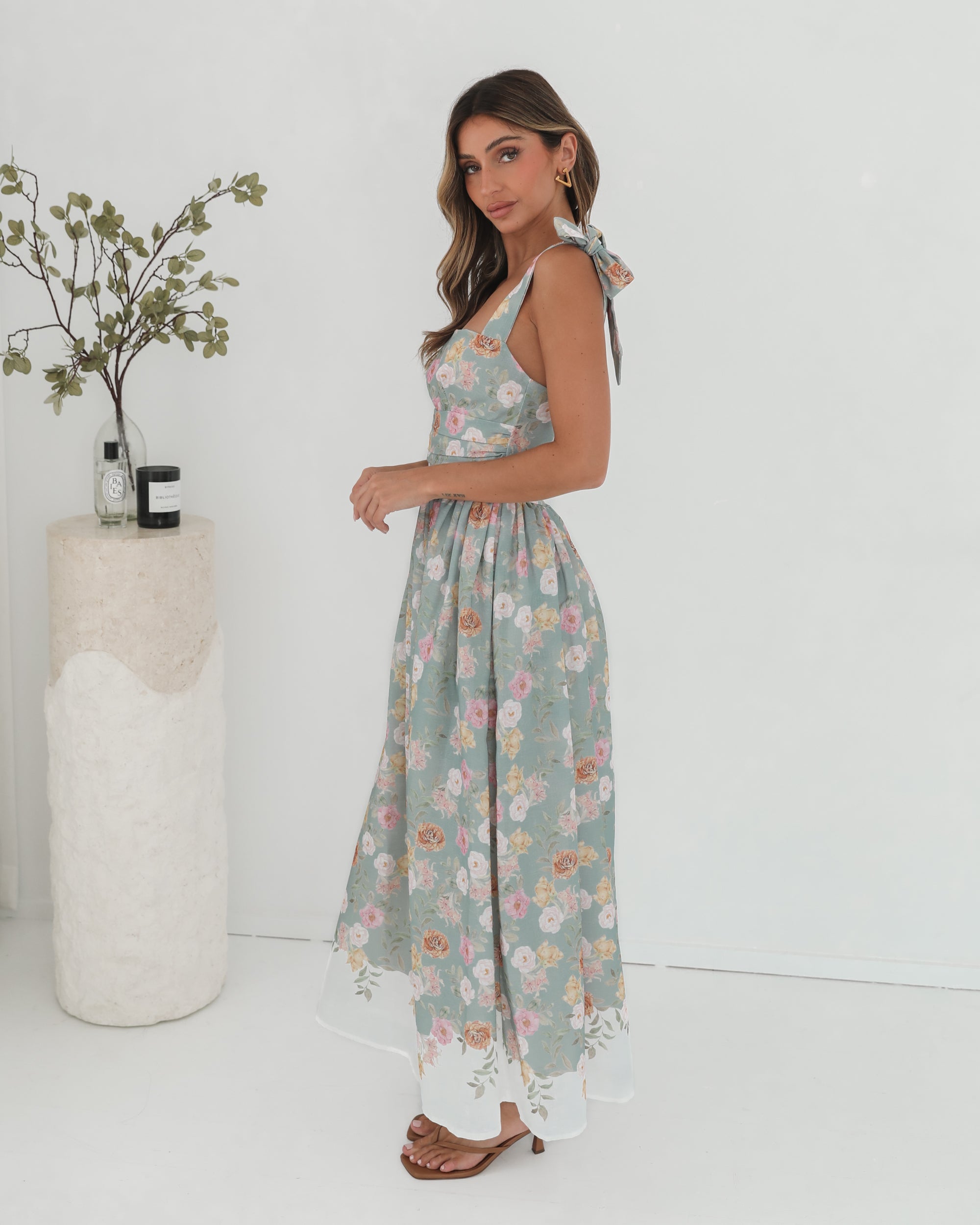 A woman standing in a room with a white background, wearing a mint floral dress with bow straps and a square neckline.