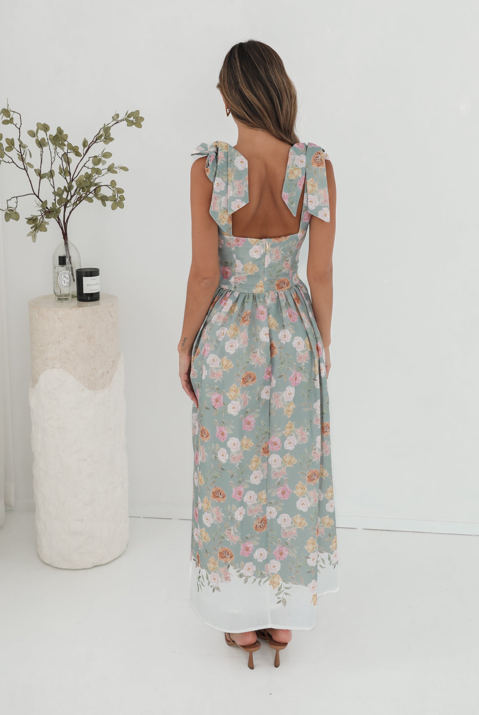 A woman standing in a room with a white background, wearing a mint floral dress with bow straps and a square neckline.