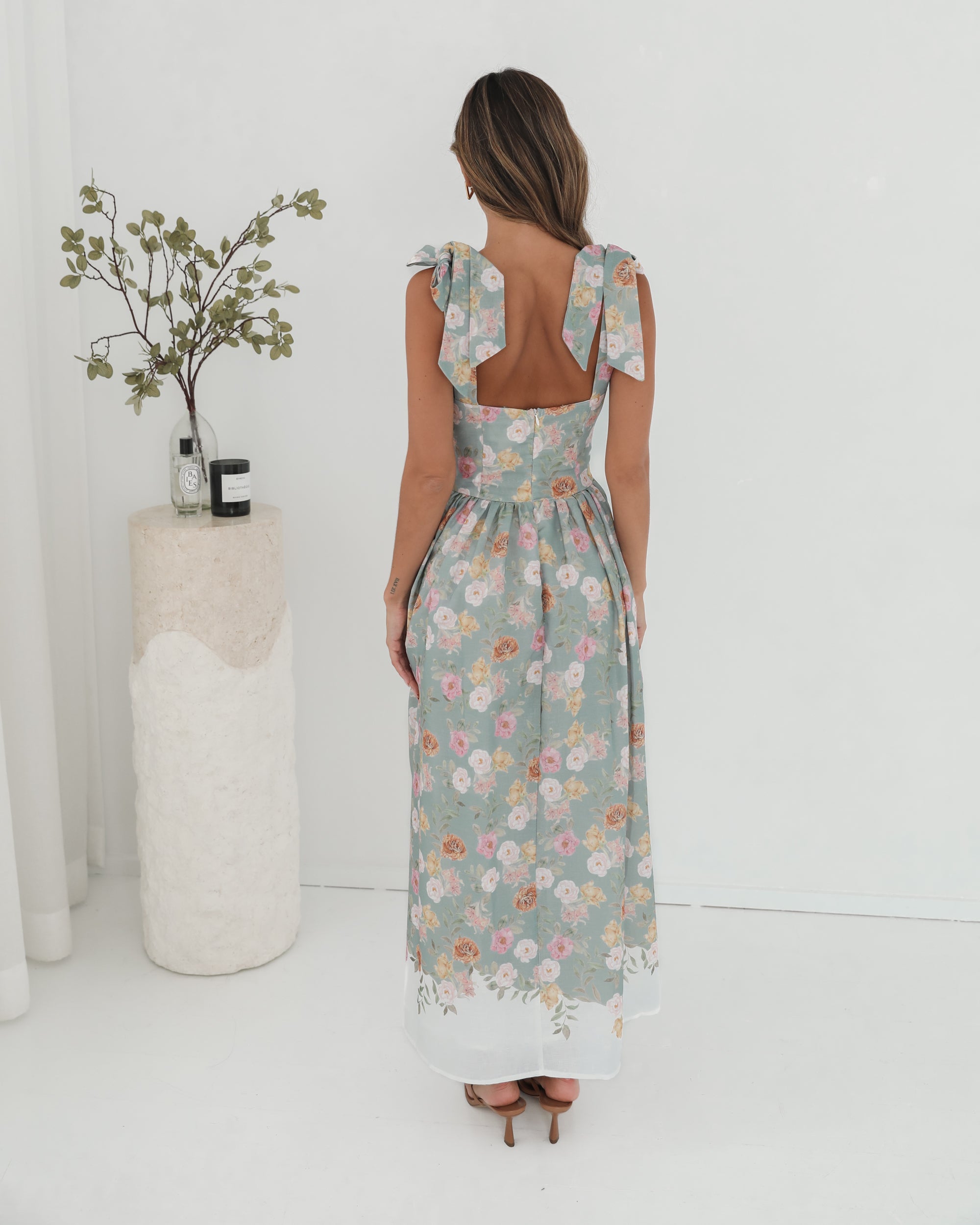 A woman standing in a room with a white background, wearing a mint floral dress with bow straps and a square neckline.