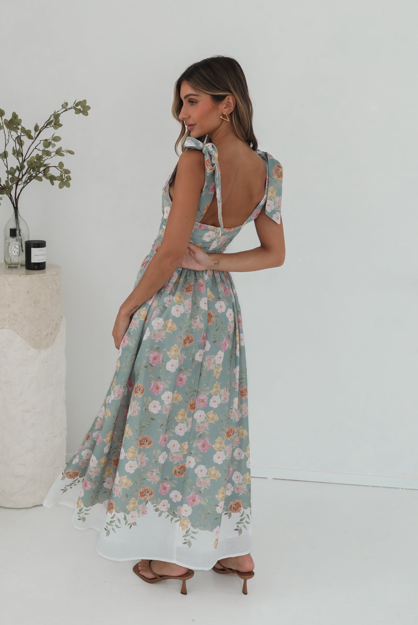 A woman standing in a room with a white background, wearing a mint floral dress with bow straps and a square neckline.
