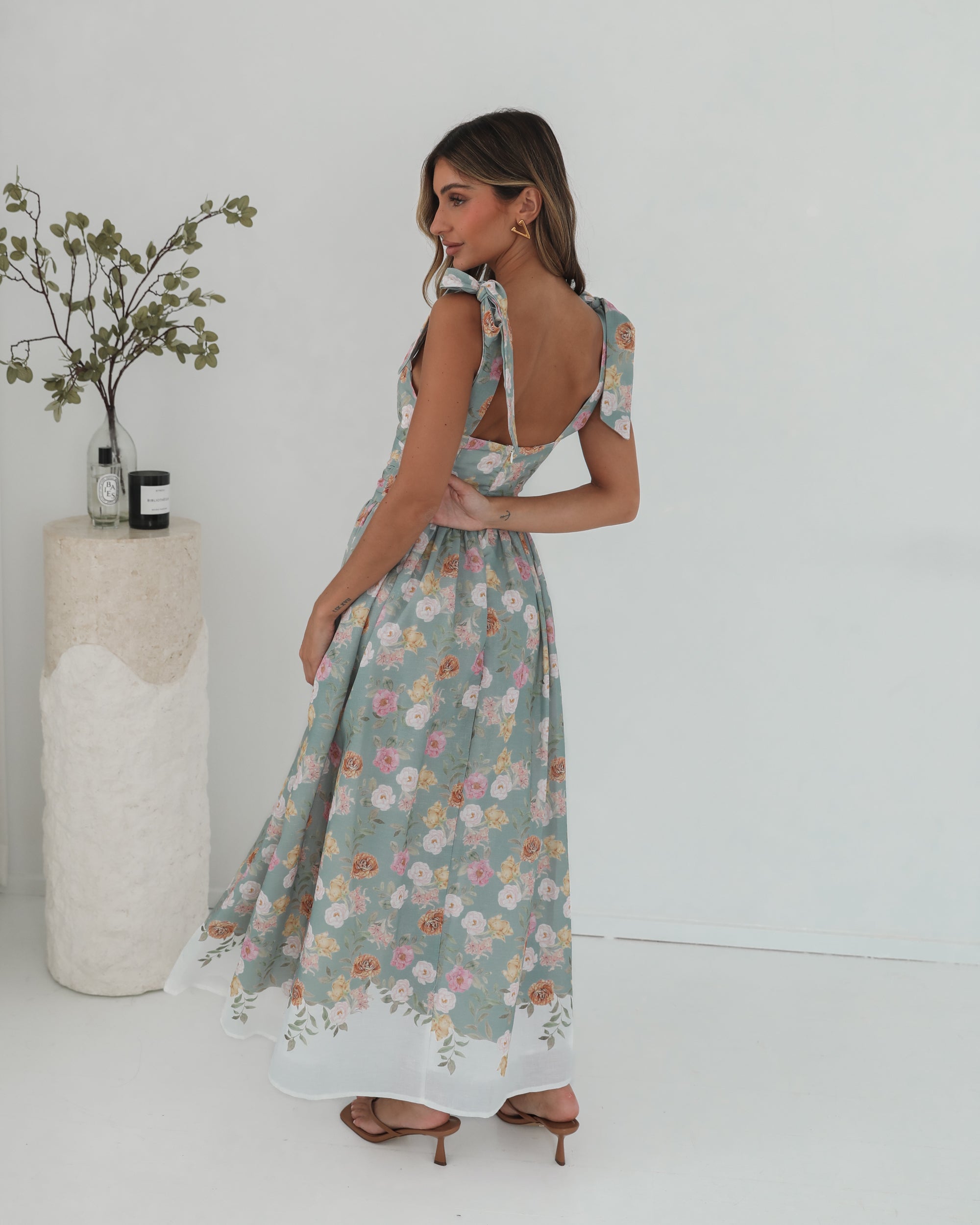 A woman standing in a room with a white background, wearing a mint floral dress with bow straps and a square neckline.