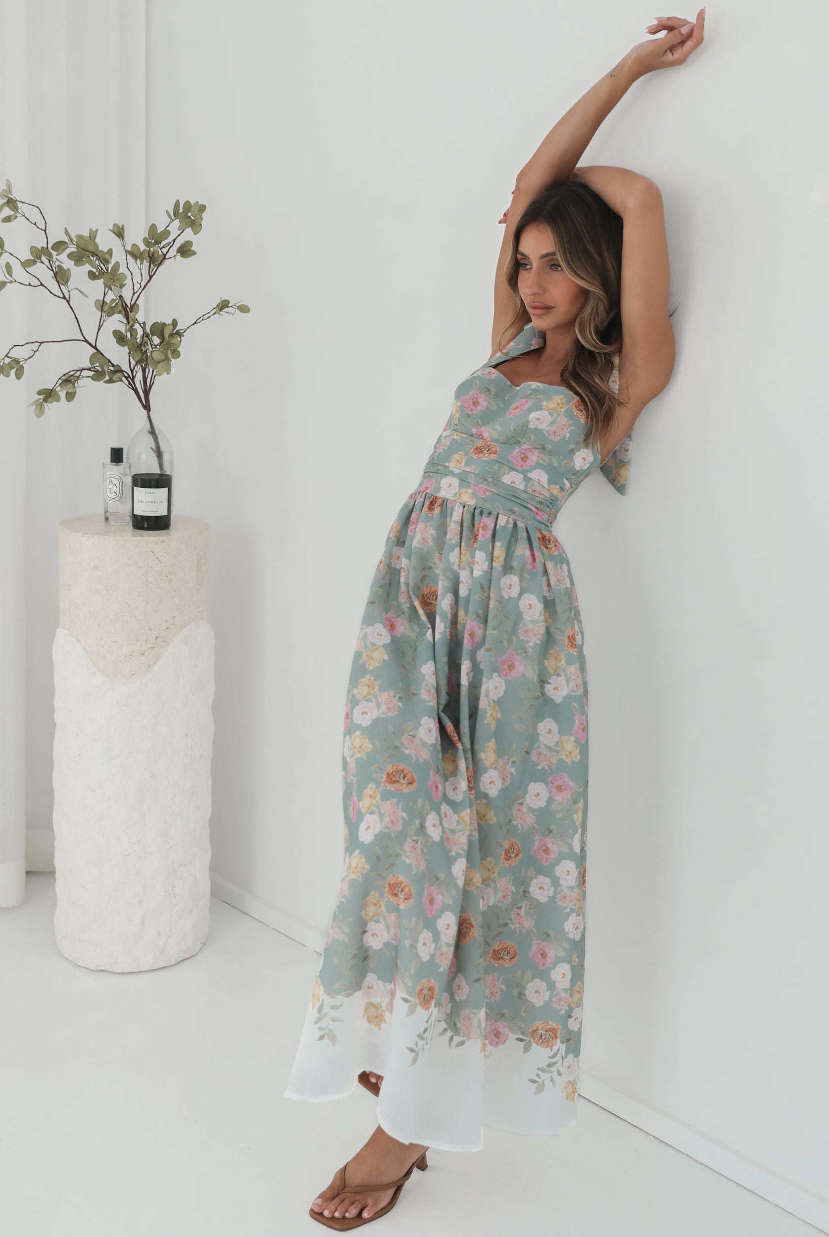 A woman standing in a room with a white background, wearing a mint floral dress with bow straps and a square neckline.
