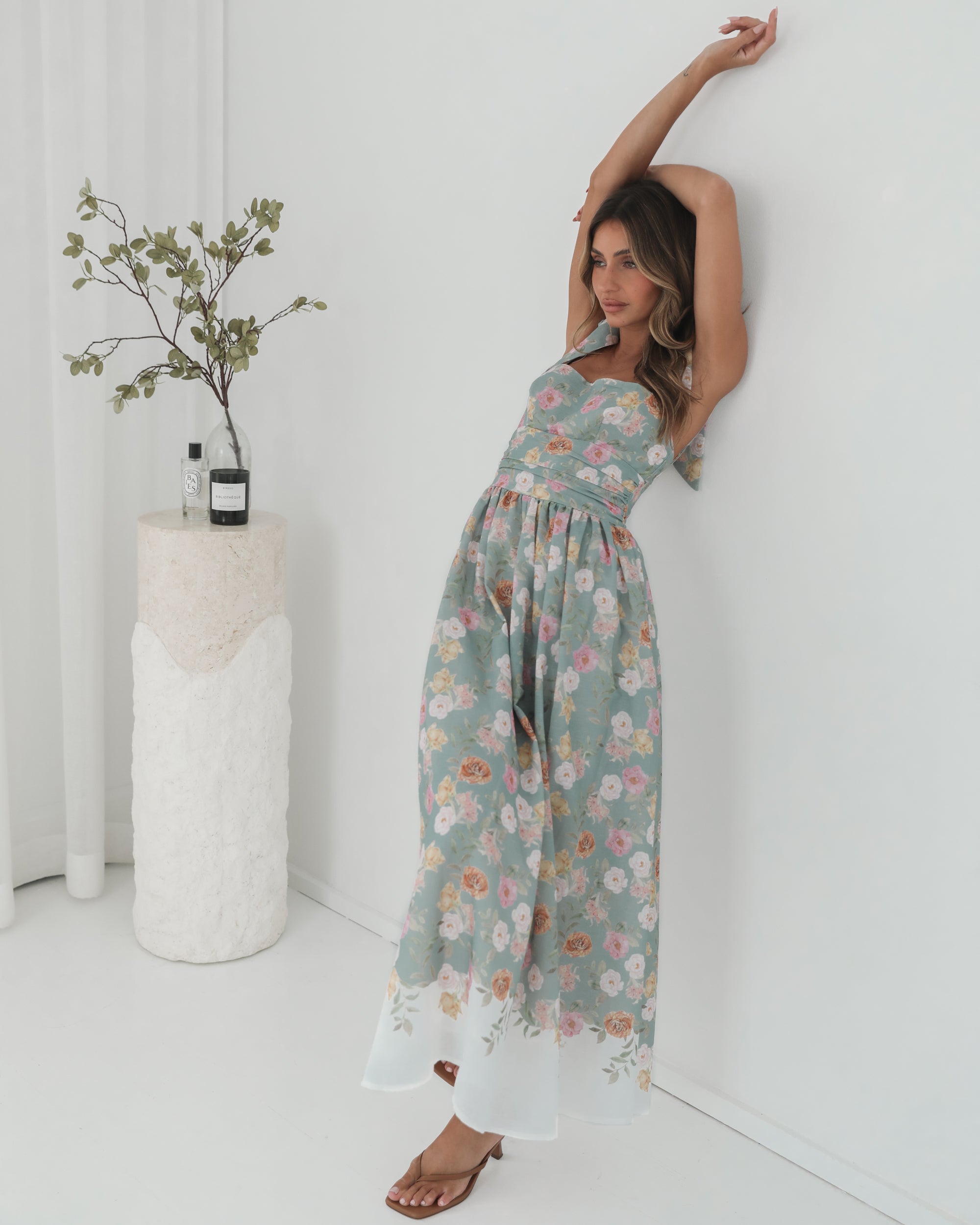 A woman standing in a room with a white background, wearing a mint floral dress with bow straps and a square neckline.