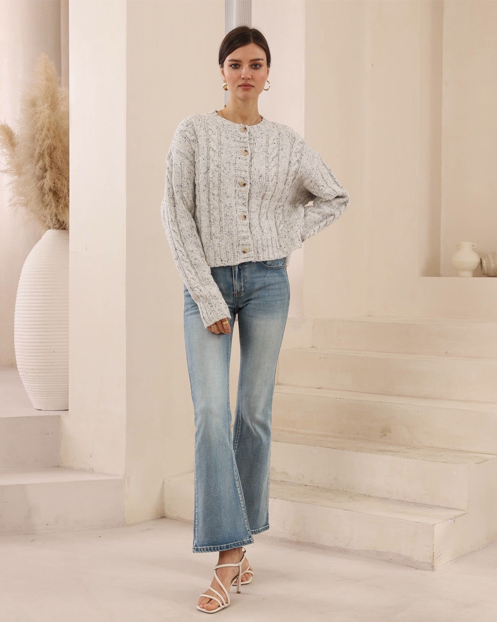 Woman wearing a light gray cardigan and blue jeans in a neutral setting