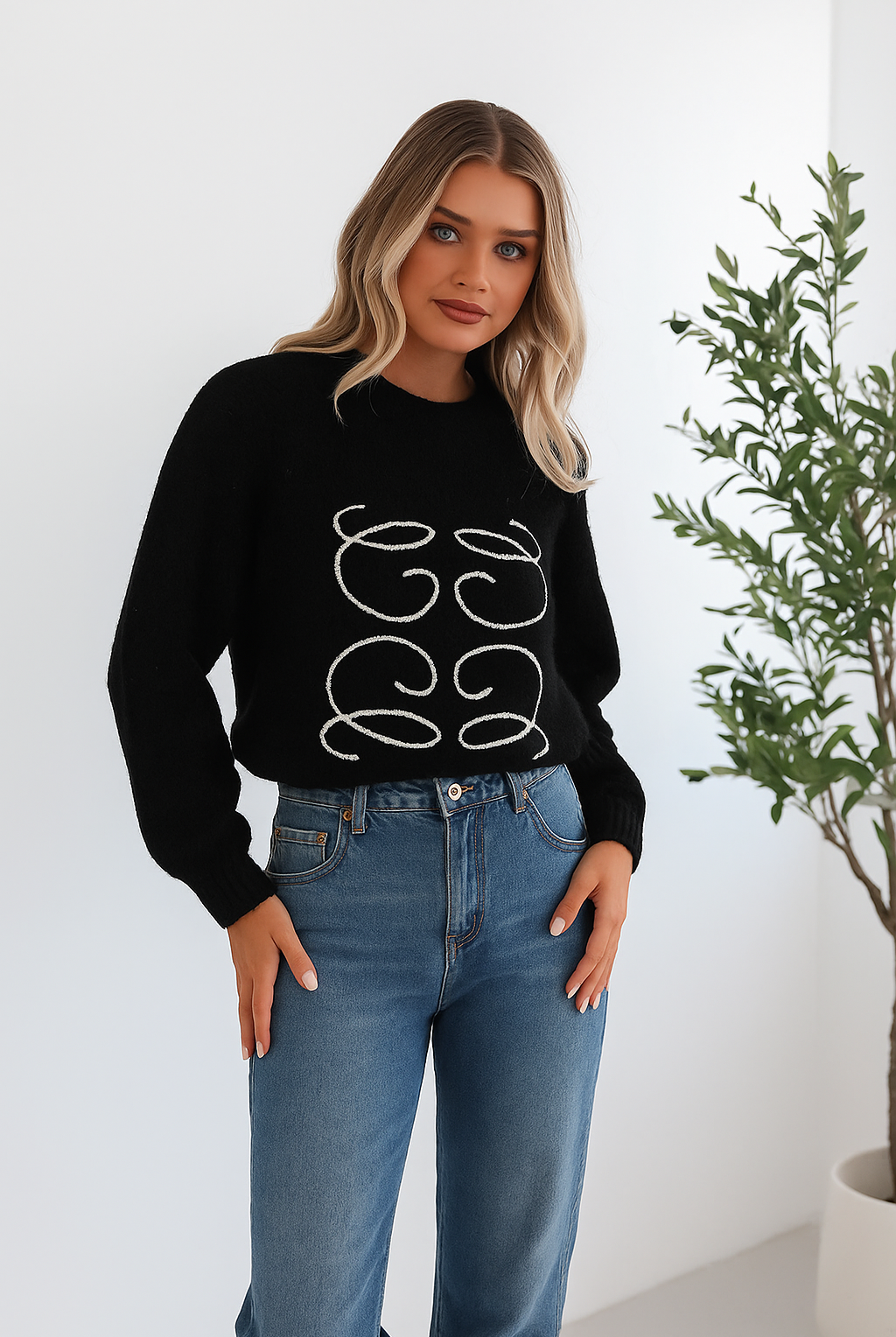 A woman with long, wavy blonde hair is standing indoors near a potted plant, wearing a black sweater with a white abstract design on the front and high-waisted blue jeans. She is looking at the camera with a neutral expression and has one hand resting on her hip. The background is minimalistic with white walls.