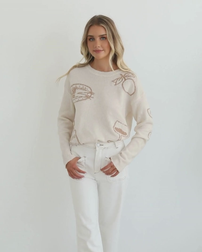 A woman wearing a cream-colored knit top with a round neckline and long sleeves, featuring a print of various items.