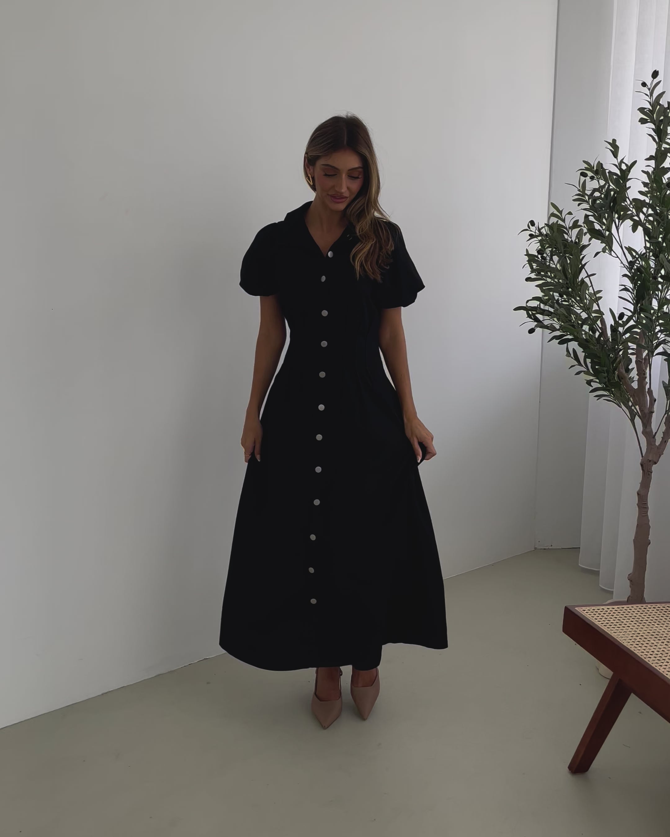 A woman standing in a room with a white background wearing a black knee-length dress with a shirt collar, short puff sleeves, and center-front buttons.