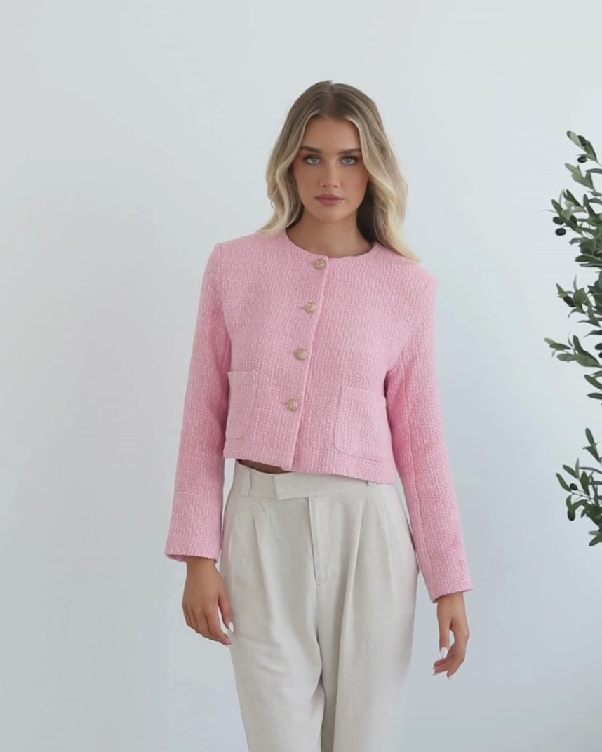 A woman wearing a pink Valdivia Jacket with long sleeves, round neckline, and center-front buttons. The jacket has a crop length and she is paired with light-colored trousers.