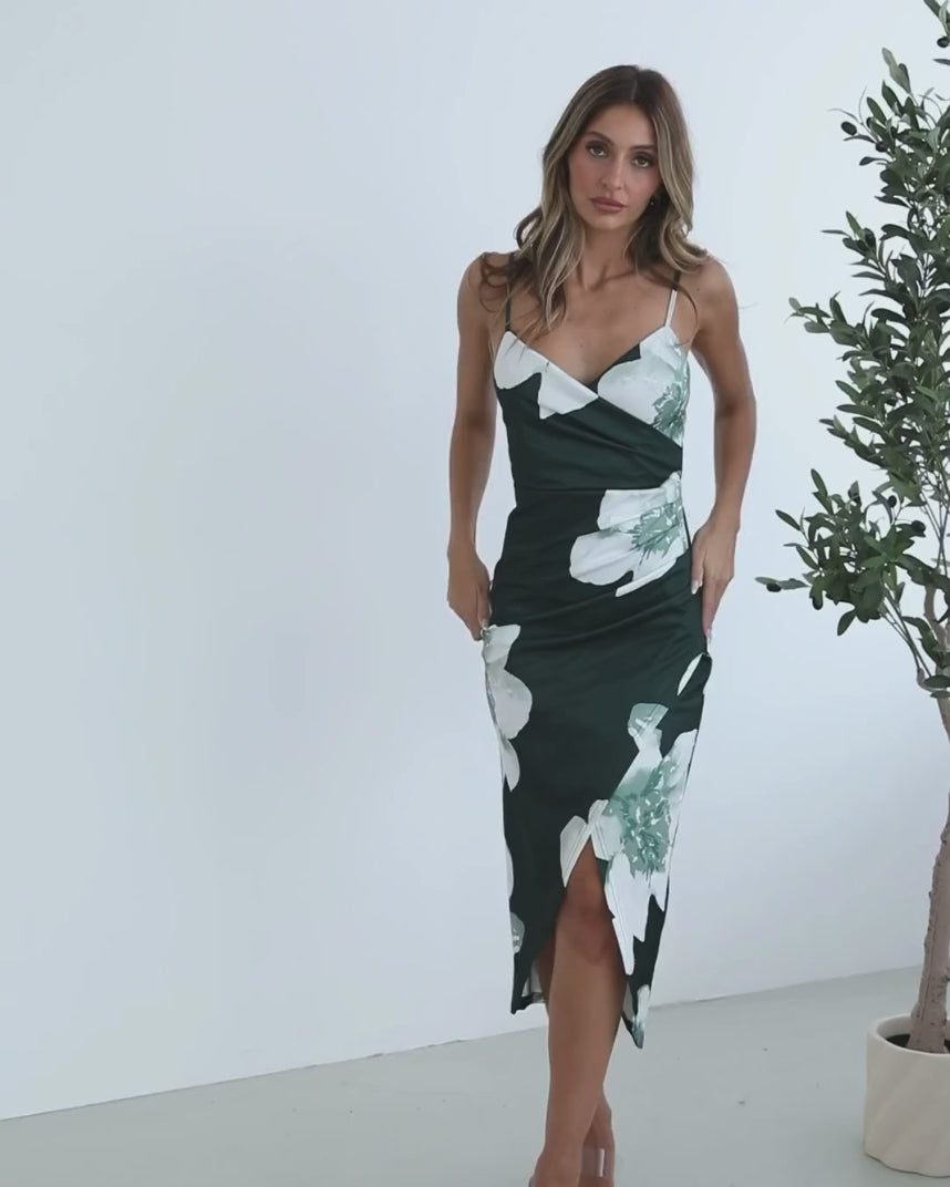 A woman wearing an emerald floral dress with spaghetti straps, a v-neckline, and an overlap skirt with a high-low hemline. She is standing next to a potted plant.