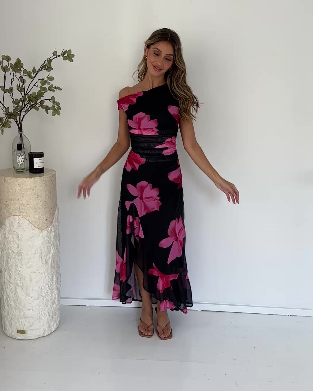 A woman standing in a white room wearing a black floral maxi dress with asymmetric neckline and ruffled skirt.