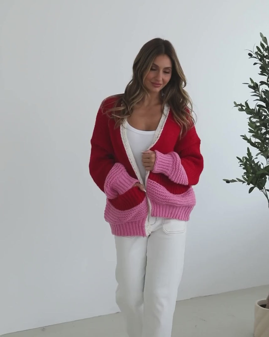 A woman sitting on a chair wearing a red, pink, and white striped cardigan with long sleeves and cuffs, paired with a white top and white pants.