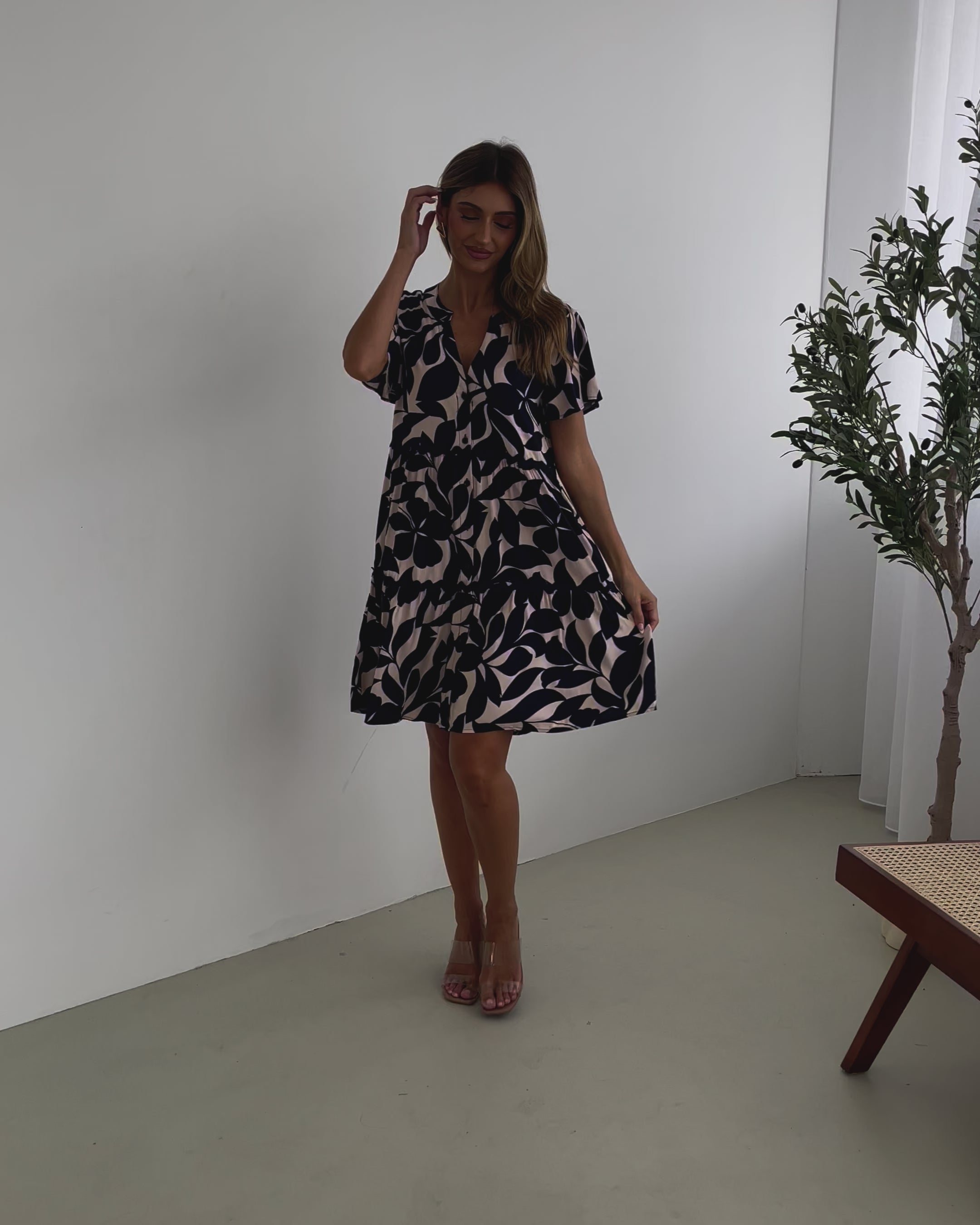A woman standing in front of a plant, wearing a beige dress with navy-tone leaf prints, short flutter sleeves, and a v-neckline.