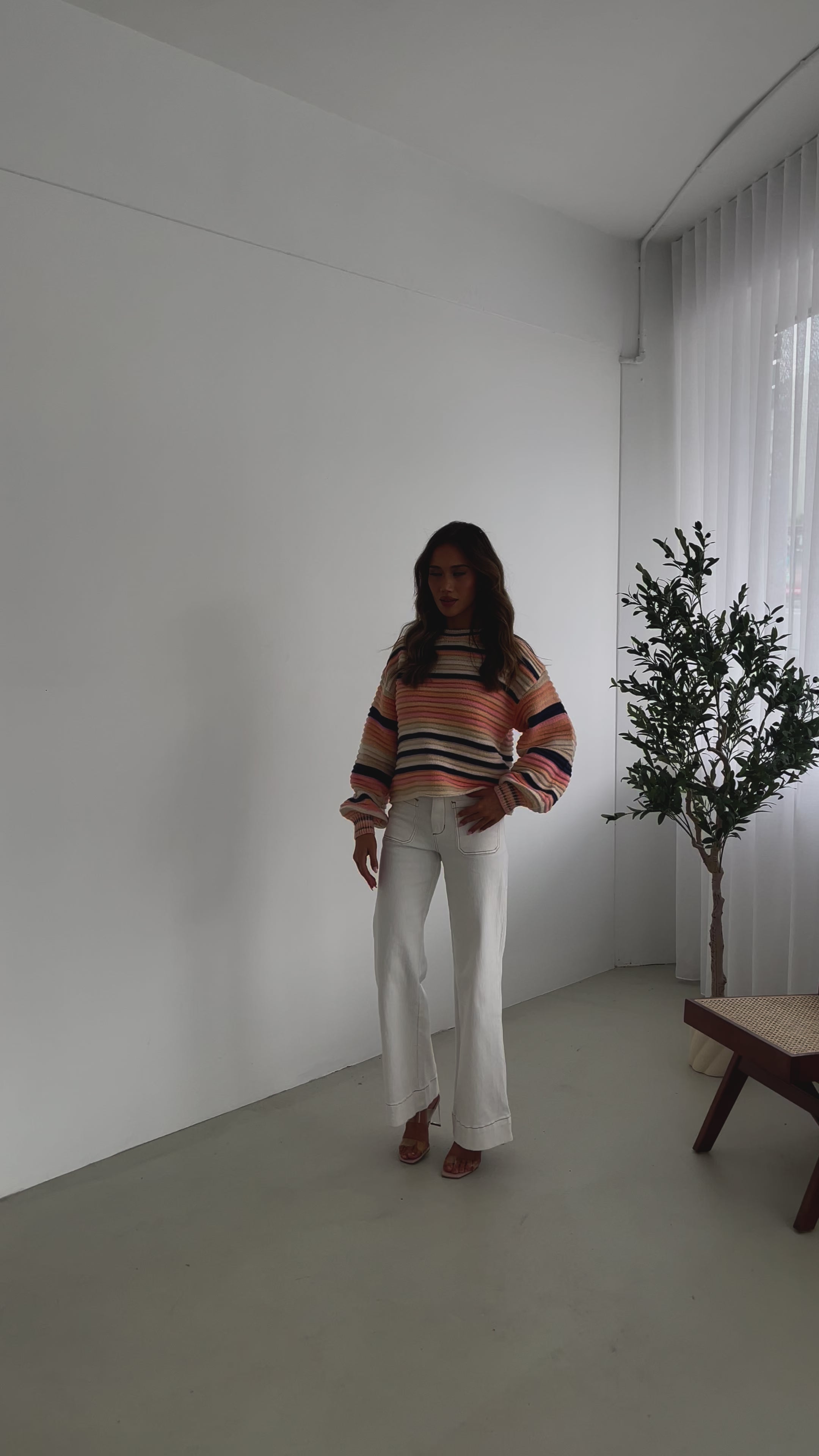 A woman sitting in a chair wearing a striped peach, pink, blue, and black knit top with a round neckline and long sleeves, paired with white pants.