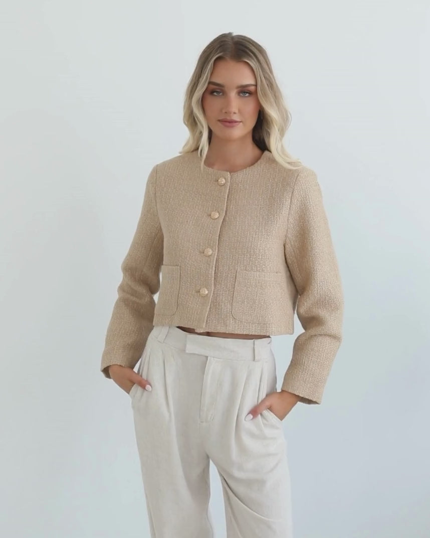 A woman wearing a beige Jacket with long sleeves, round neckline, and center-front buttons. The jacket has a crop length and she is paired with light-colored trousers.