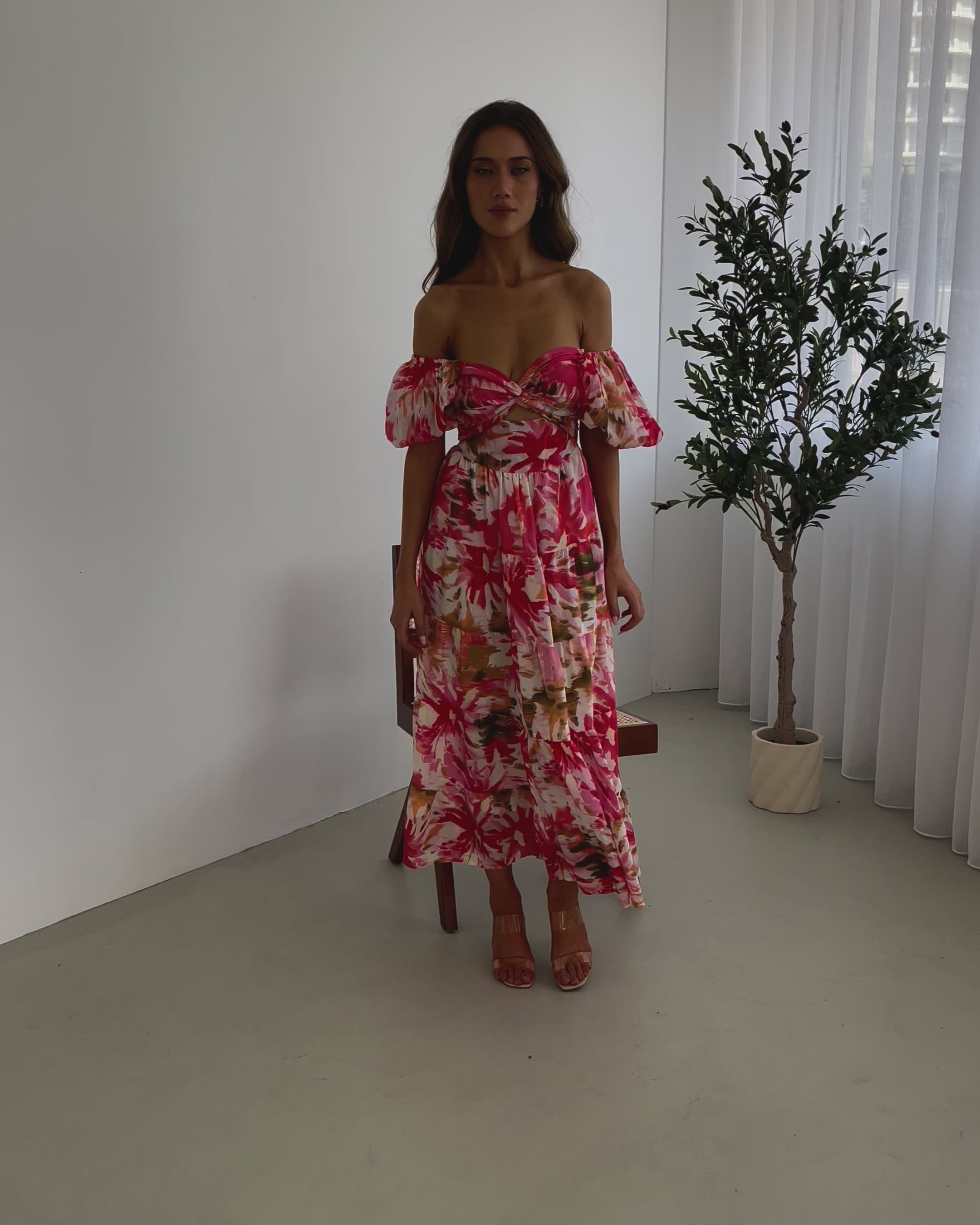 A woman standing next to a chair, wearing a floral print maxi dress with a sweetheart neckline and short balloon sleeves. She is posing in a room with a large window and a plant in the background.