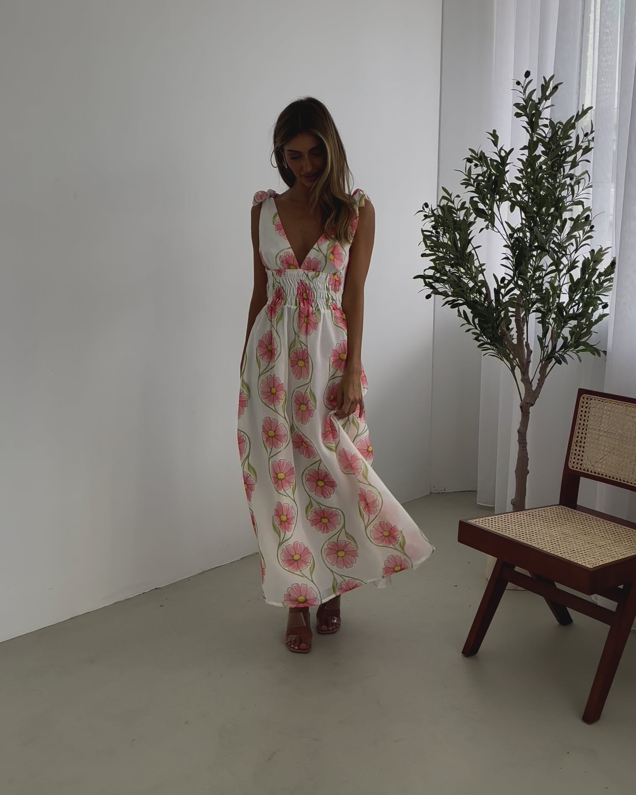 A woman standing in a room with a white floral maxi dress with bow straps and a plunge neckline.