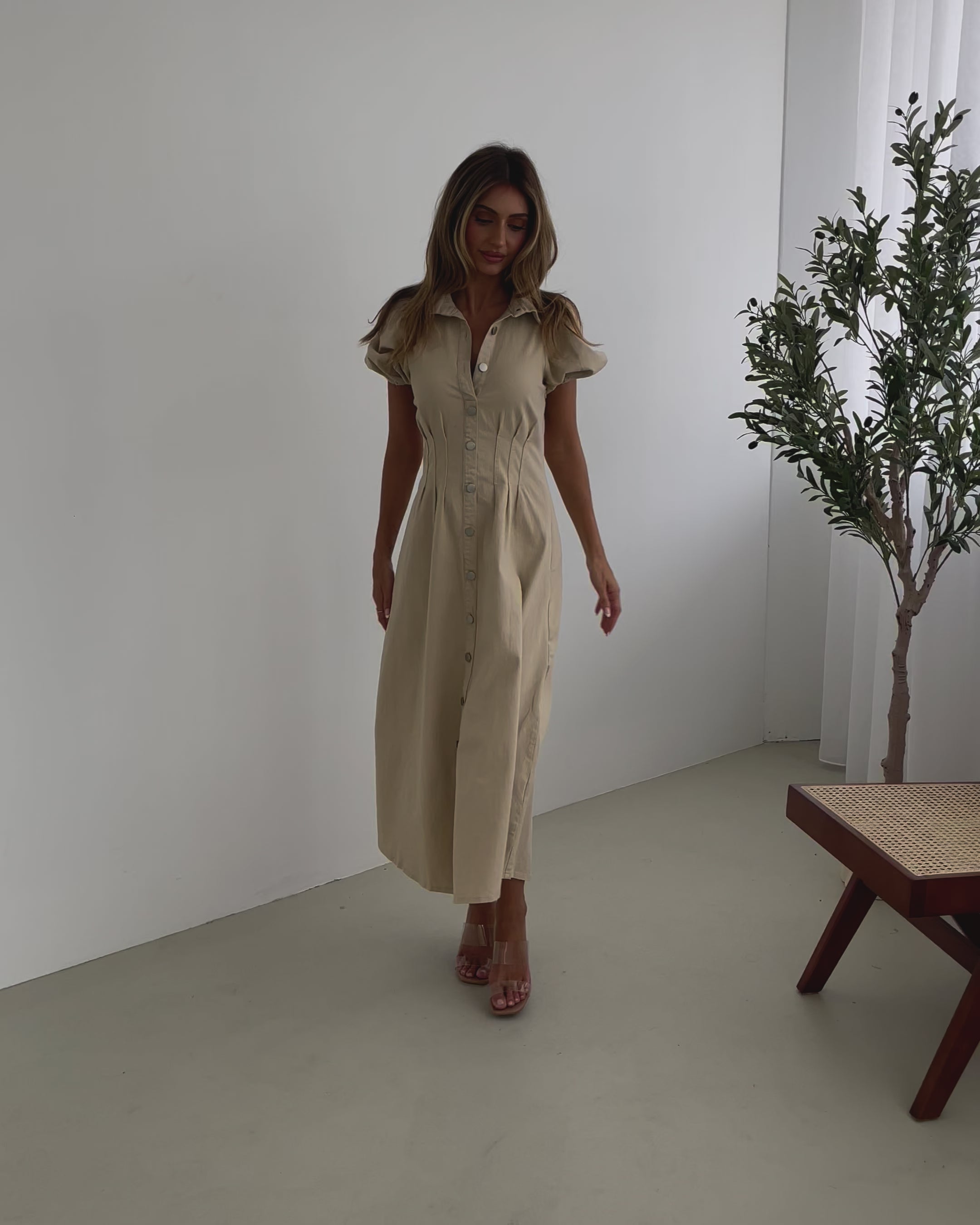 A woman standing in a room with a beige dress with a shirt collar, short puff sleeves, and a-line skirt with a midaxi length hem.