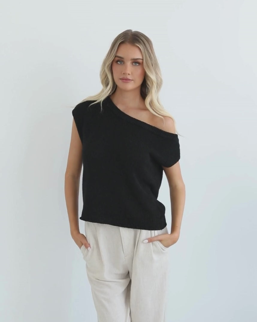 A female model wearing a black knit top with a one side off shoulder design, paired with light-colored trousers.