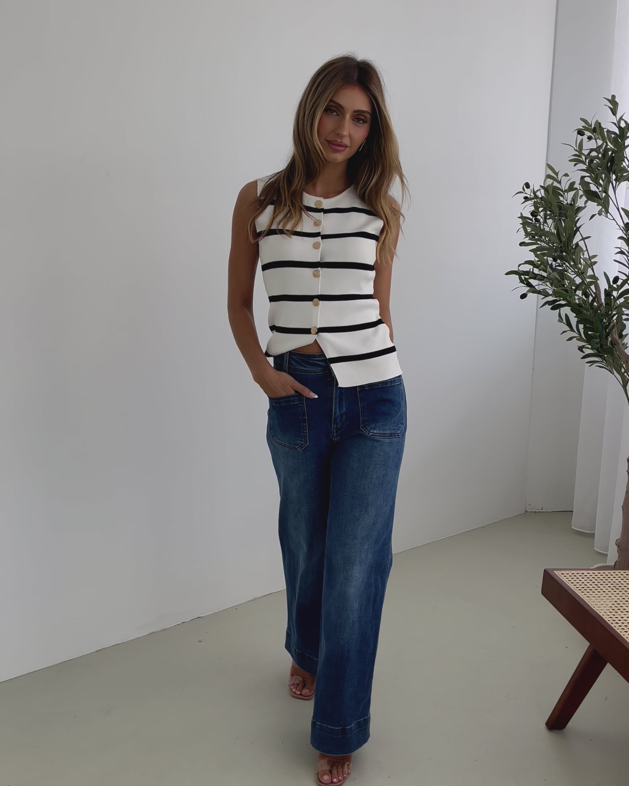 A woman standing in a room with light decor, wearing a striped top and mid-blue Yzabel Jeans with a high waist fit and wide leg silhouette.