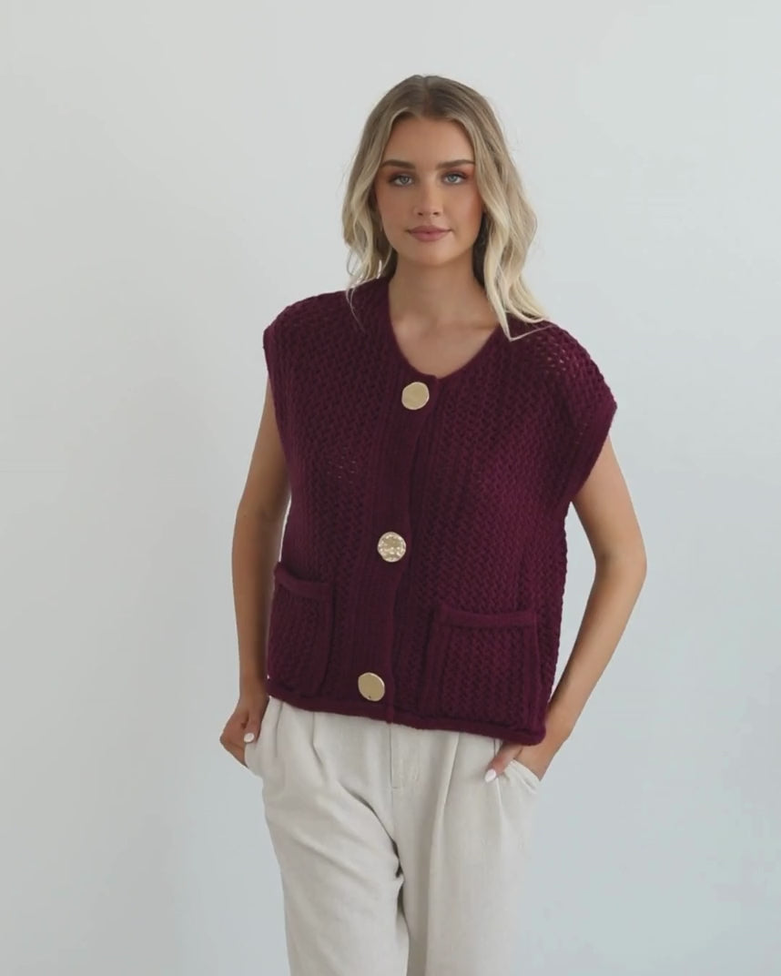 A woman wearing a sleeveless wine-colored knit top with round neckline and center-front buttons, paired with light-colored pants.