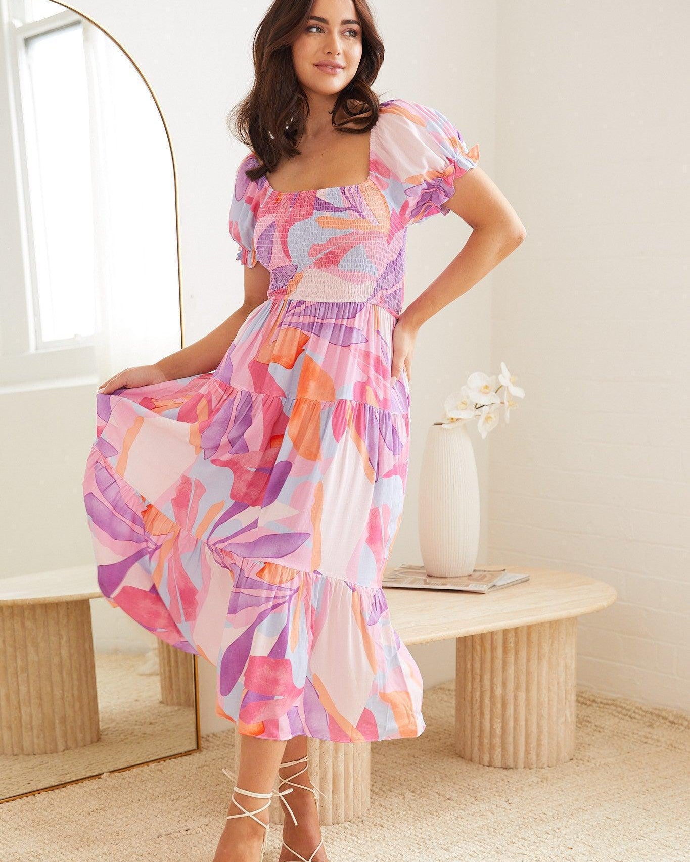 A woman wearing a colorful, abstract-patterned midi dress in shades of pink, purple, and orange. The dress features puff sleeves, a smocked bodice, and a tiered skirt. She poses indoors near a modern wooden table with a white vase holding white flowers, and a large arched mirror in the background.