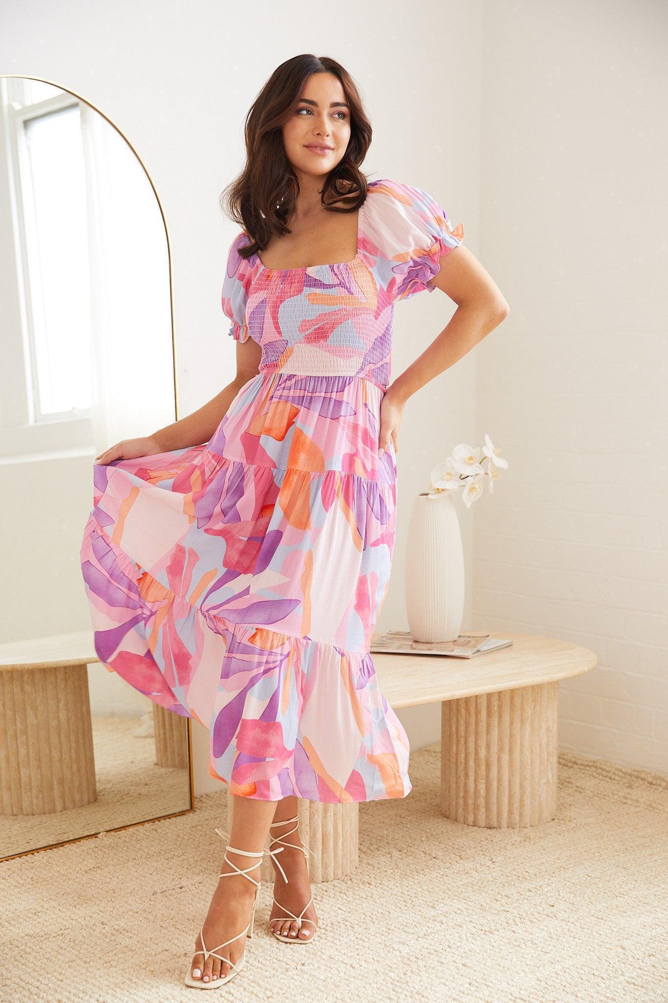 A woman wearing a colorful, abstract-patterned midi dress in shades of pink, purple, and orange. The dress features puff sleeves, a smocked bodice, and a tiered skirt. She poses indoors near a modern wooden table with a white vase holding white flowers, and a large arched mirror in the background.