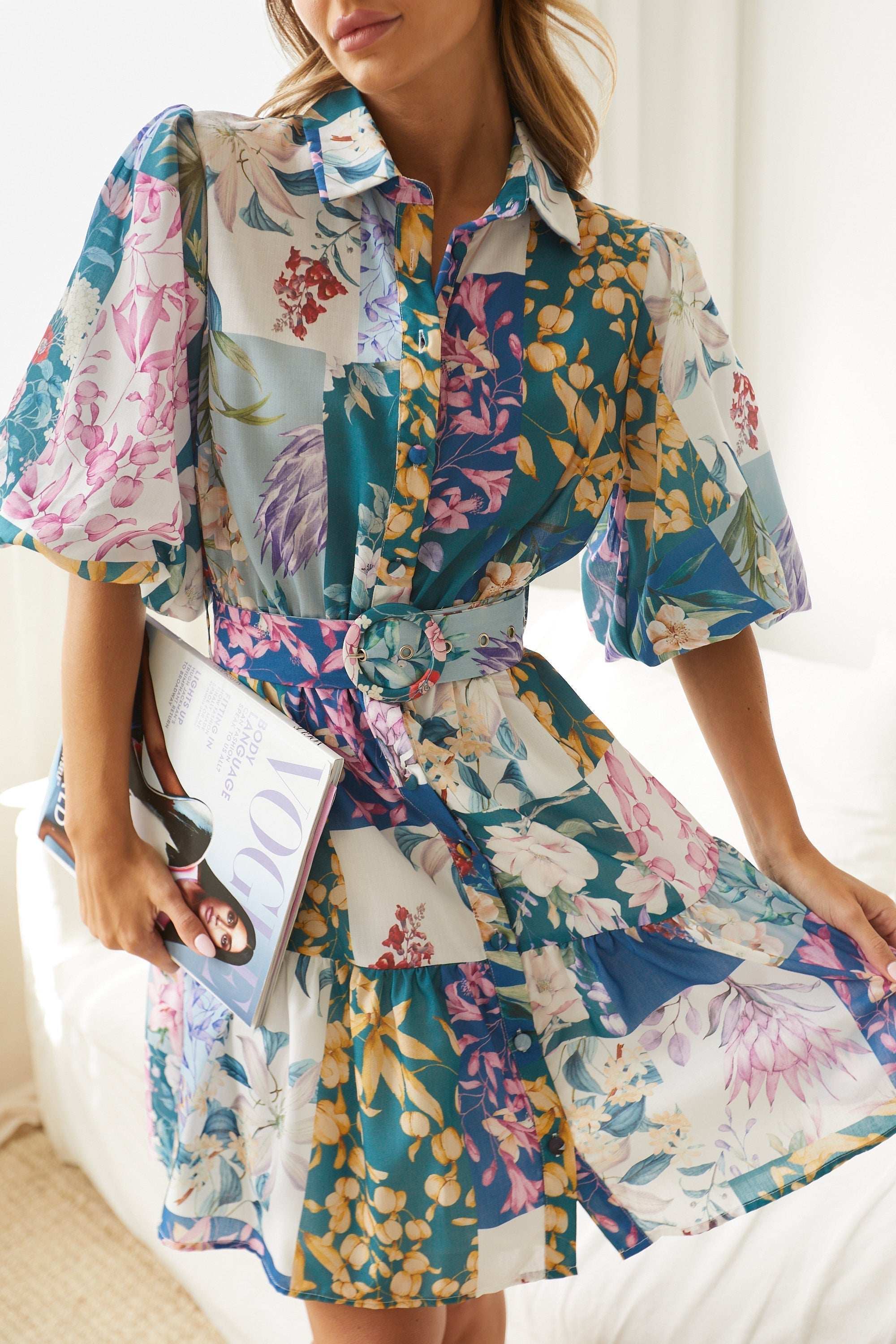A woman wearing a short, multicolored floral dress with puff sleeves and a belted waist. The dress features a patchwork design with vibrant flower patterns in shades of pink, blue, yellow, and green. She is standing indoors in front of a white couch, looking to the side with a gentle smile.