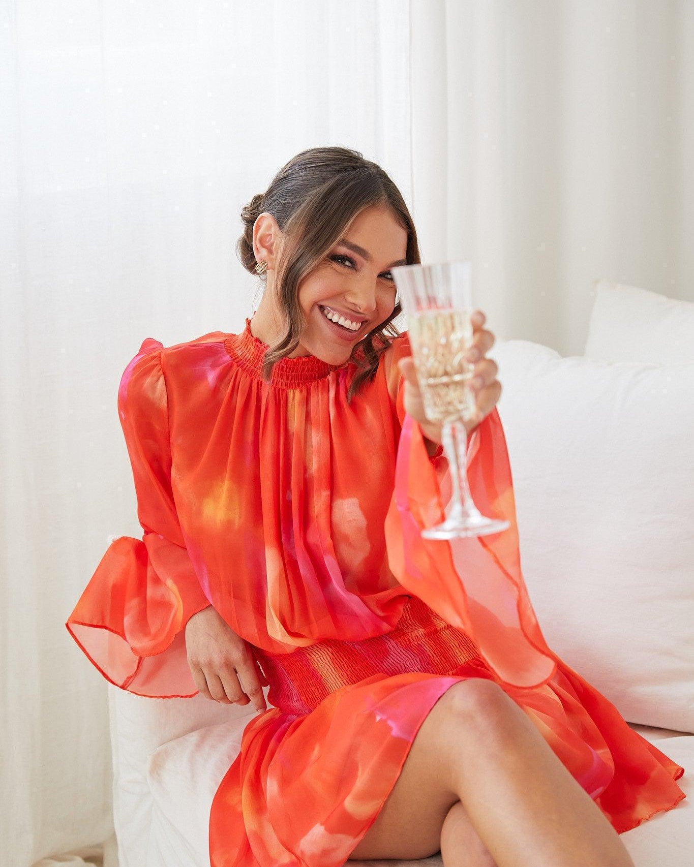 A woman sitting on a white couch, holding a glass of champagne, wearing an orange base dress with a red and purple print, featuring a high neckline and long trumpet sleeves.
