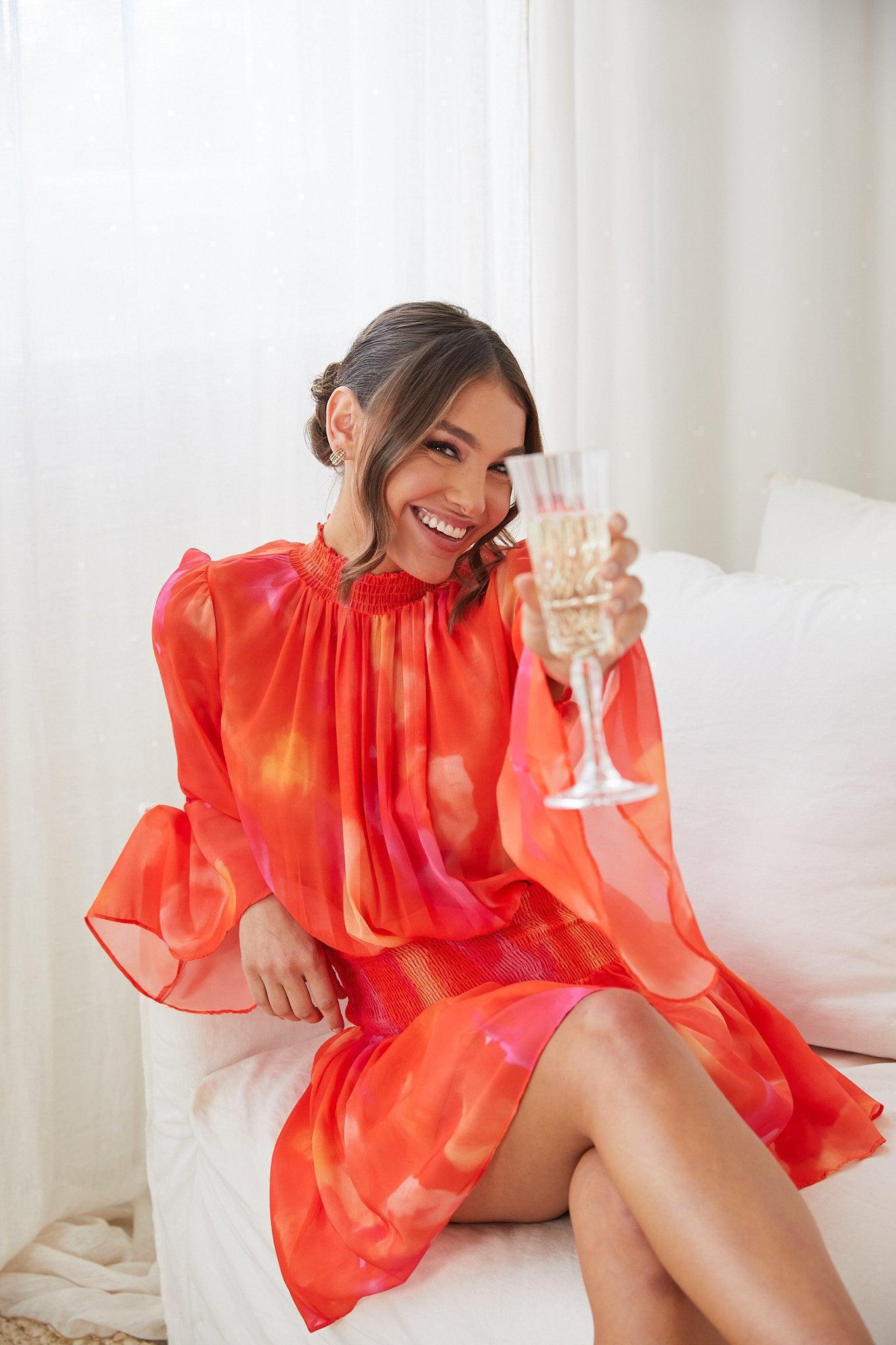 A woman sitting on a white couch, holding a glass of champagne, wearing an orange base dress with a red and purple print, featuring a high neckline and long trumpet sleeves.