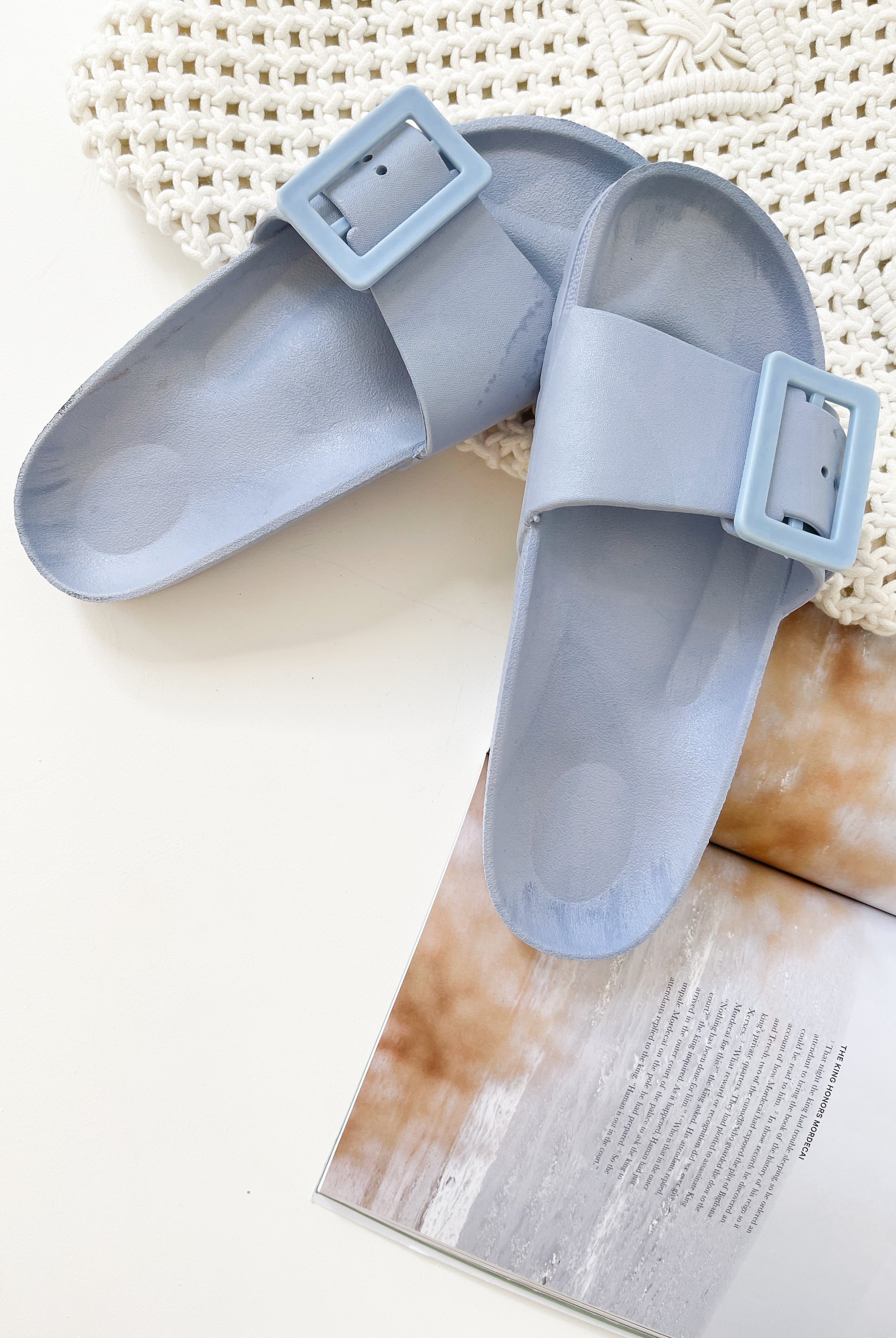 A pair of black summer slides with thick straps, displayed on a flat surface with a knitted texture in the background.