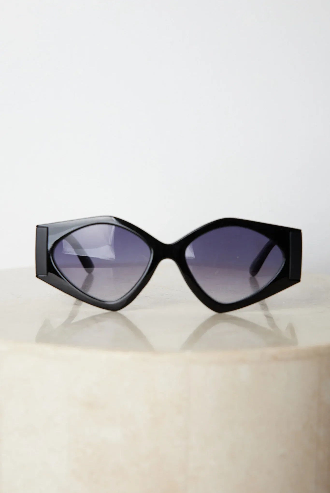 Black cat-eye sunglasses with thick frames and dark tinted lenses, displayed on a light beige surface against a plain white background.