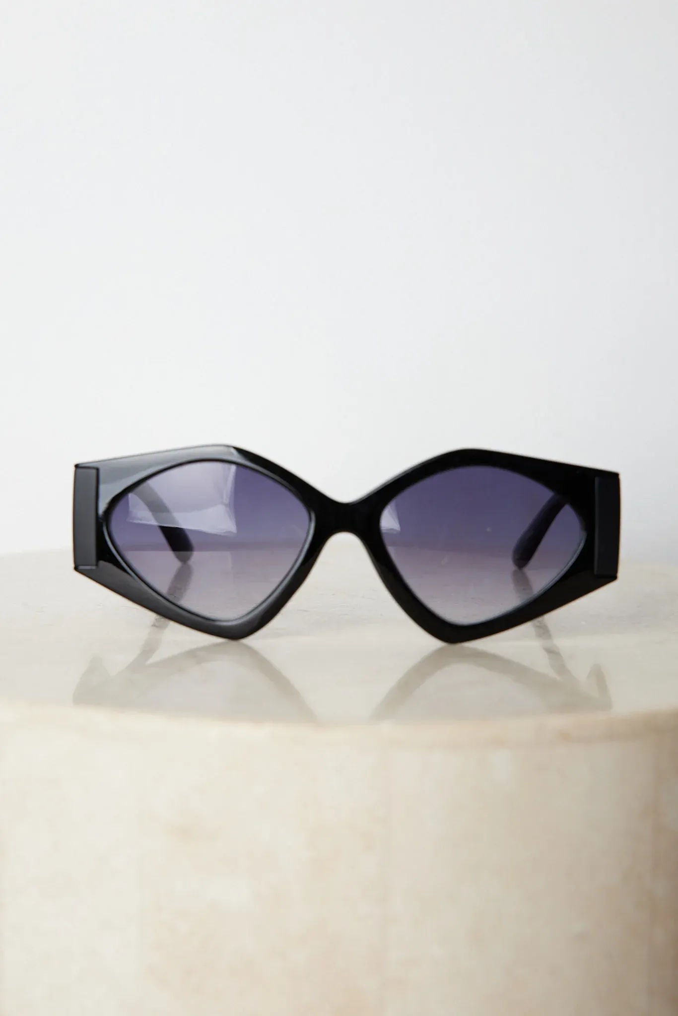 Black cat-eye sunglasses with thick frames and dark tinted lenses, displayed on a light beige surface against a plain white background.