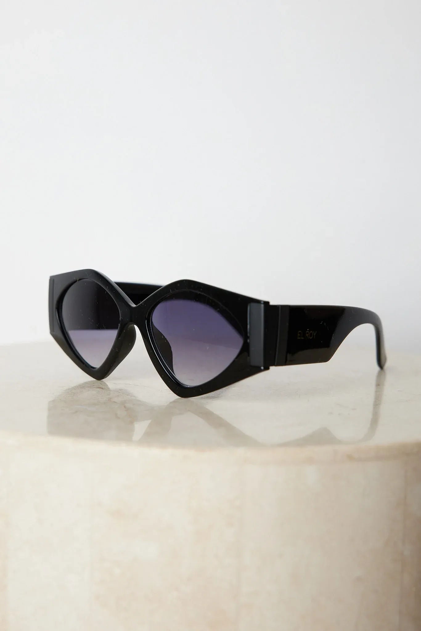 Black cat-eye sunglasses with thick frames and dark tinted lenses, displayed on a light beige surface against a plain white background.