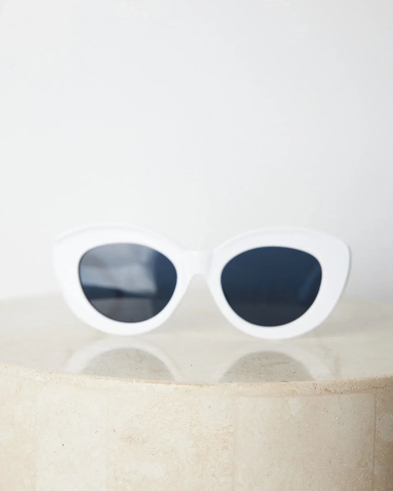 White cat-eye sunglasses with dark lenses displayed on a light beige marble surface against a plain white background.