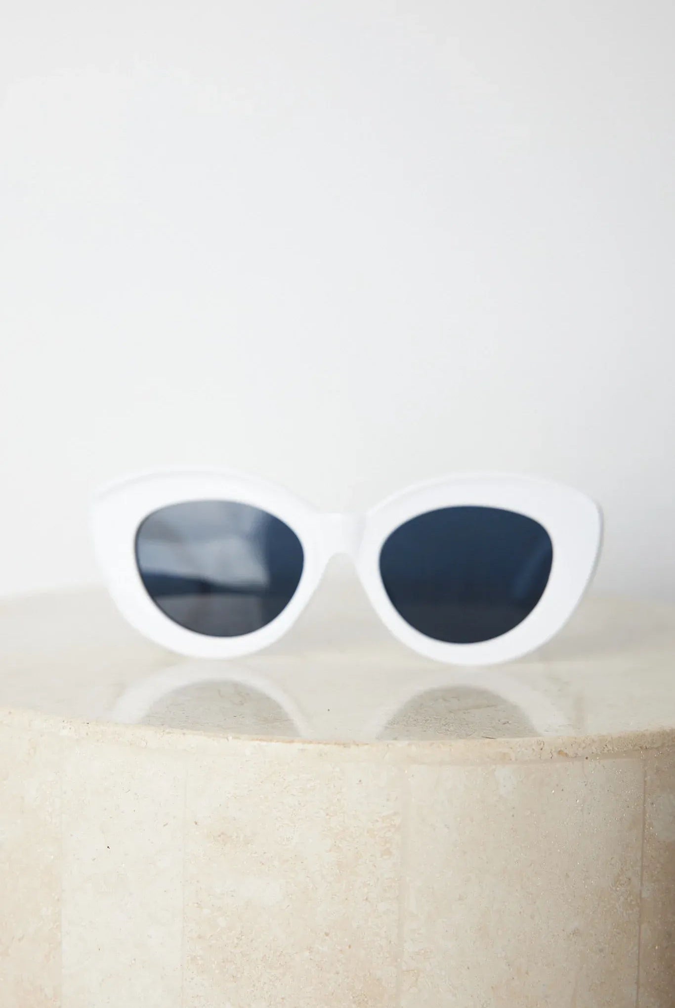 White cat-eye sunglasses with dark lenses displayed on a light beige marble surface against a plain white background.