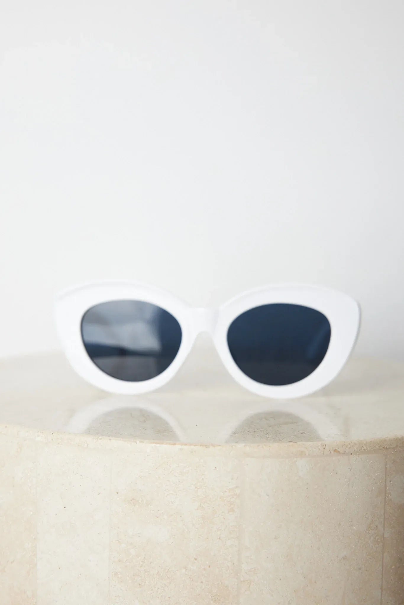 White cat-eye sunglasses with dark lenses displayed on a light beige marble surface against a plain white background.