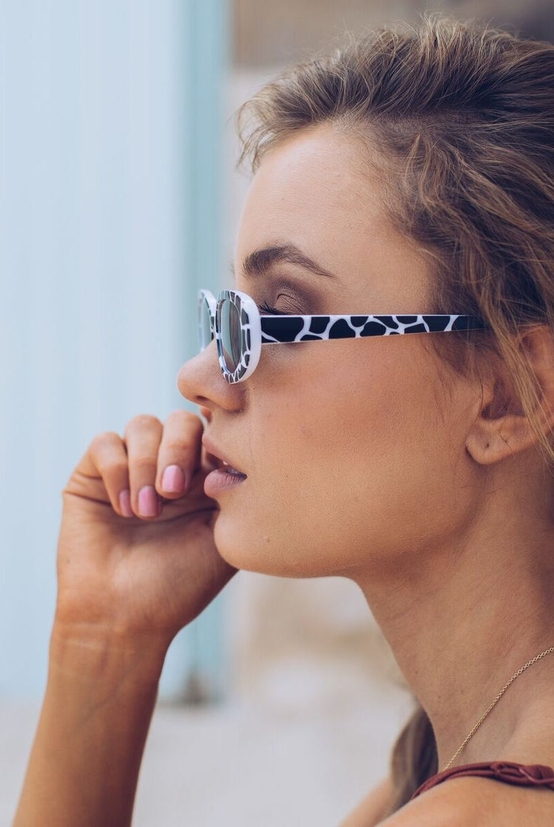 Woman wearing narrow oval-shaped sunglasses with a bold black and white animal print frame, looking over her shoulder with a confident expression. She has wavy blonde hair and is dressed in a rust-colored spaghetti strap top, sitting against a soft beachy backdrop with sand, a light blue door, and a stone wall.